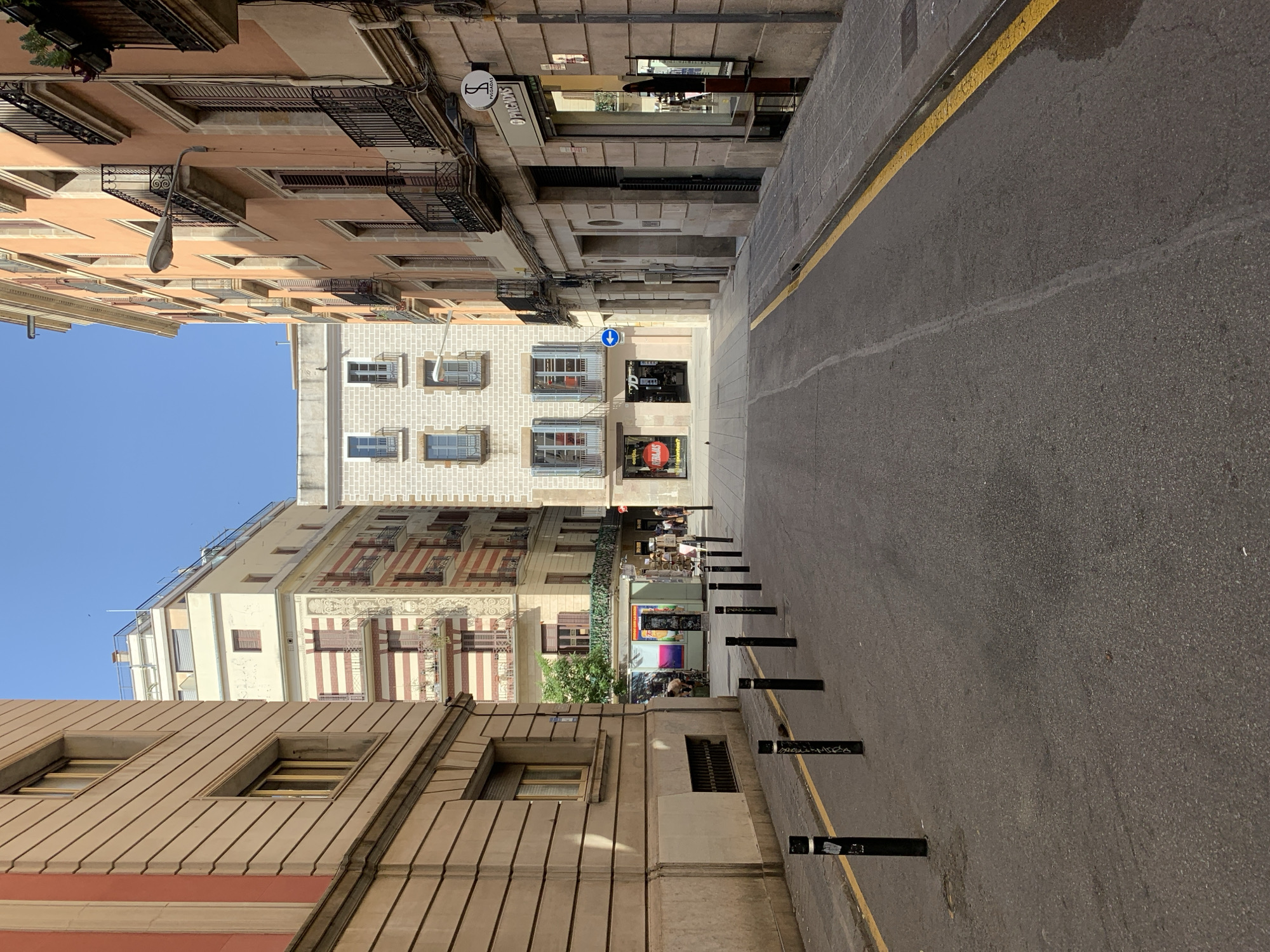 The image depicts a street scene in Barcelona, Spain, likely in the Gothic Quarter or El Born, characterized by narrow streets lined with multi-story buildings. The perspective looks down a street with buildings on both sides. On the left, a building with a beige facade and horizontal banding dominates the foreground. Further down the street, a cream-colored building with red and white striped accents is visible. On the right, a building with an orange-ochre facade features multiple balconies with black wrought-iron railings.
The street itself is paved with asphalt and marked with a yellow line along the curb. Several black bollards are positioned along the sidewalk on the left side of the street. In the distance, a small kiosk or stand with colorful displays is situated near the base of the striped building, with a few individuals visible nearby. The sky is clear and blue, indicating a sunny day. Architectural details such as windows with shutters and balconies suggest a European urban environment.