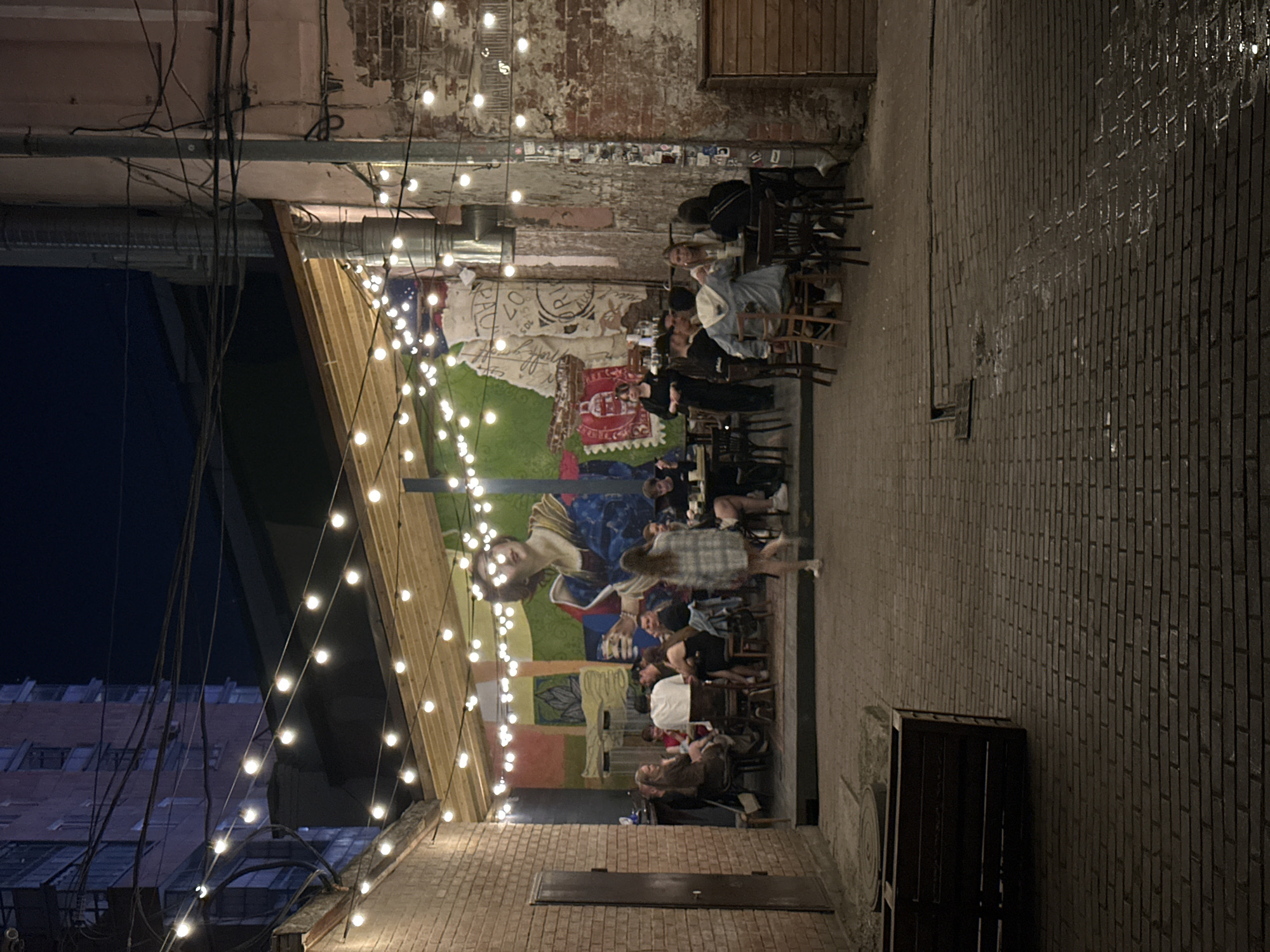 This image depicts an outdoor dining area in Perm, Russia, illuminated by string lights. A mural covers the back wall behind the tables where patrons are seated and standing, engaged in conversation and dining. The area is enclosed by brick buildings and walls, with a dark sky visible above. There is a brick pavement and a metal crate or structure on the right. The scene appears to be in a courtyard or alleyway.