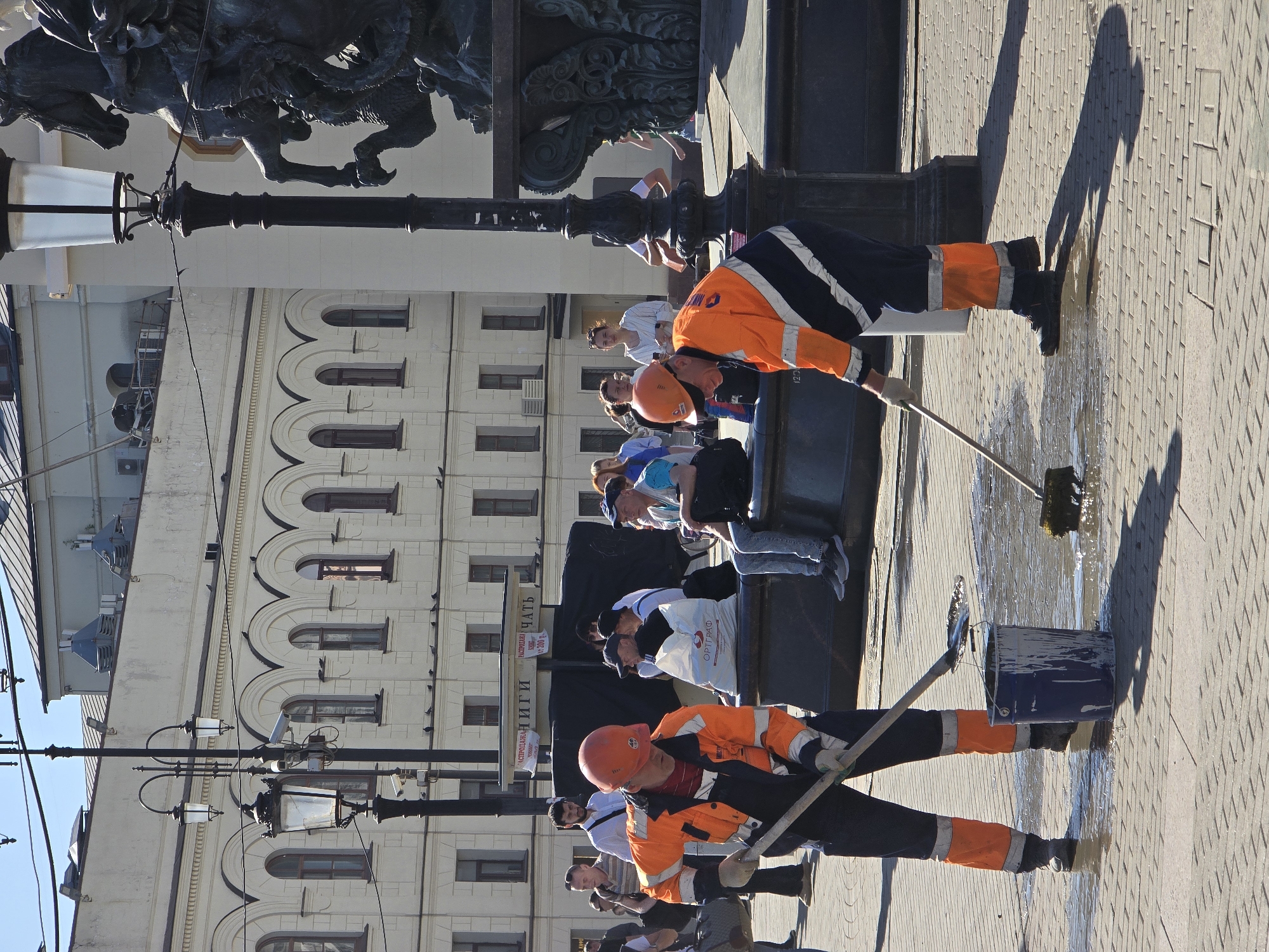 The image shows two workers wearing orange and navy uniforms and hard hats cleaning a brick-paved area in what appears to be a public space. One worker uses a long-handled tool, possibly a squeegee or brush, to clean the pavement, while a bucket sits nearby. Other people are visible in the background, including a group sitting or standing near the base of a monument or statue. In the background is a building with arched windows. Based on the architecture and the statue of St. George slaying the dragon, this scene is most likely in Manezhnaya Square in Moscow, Russia.
