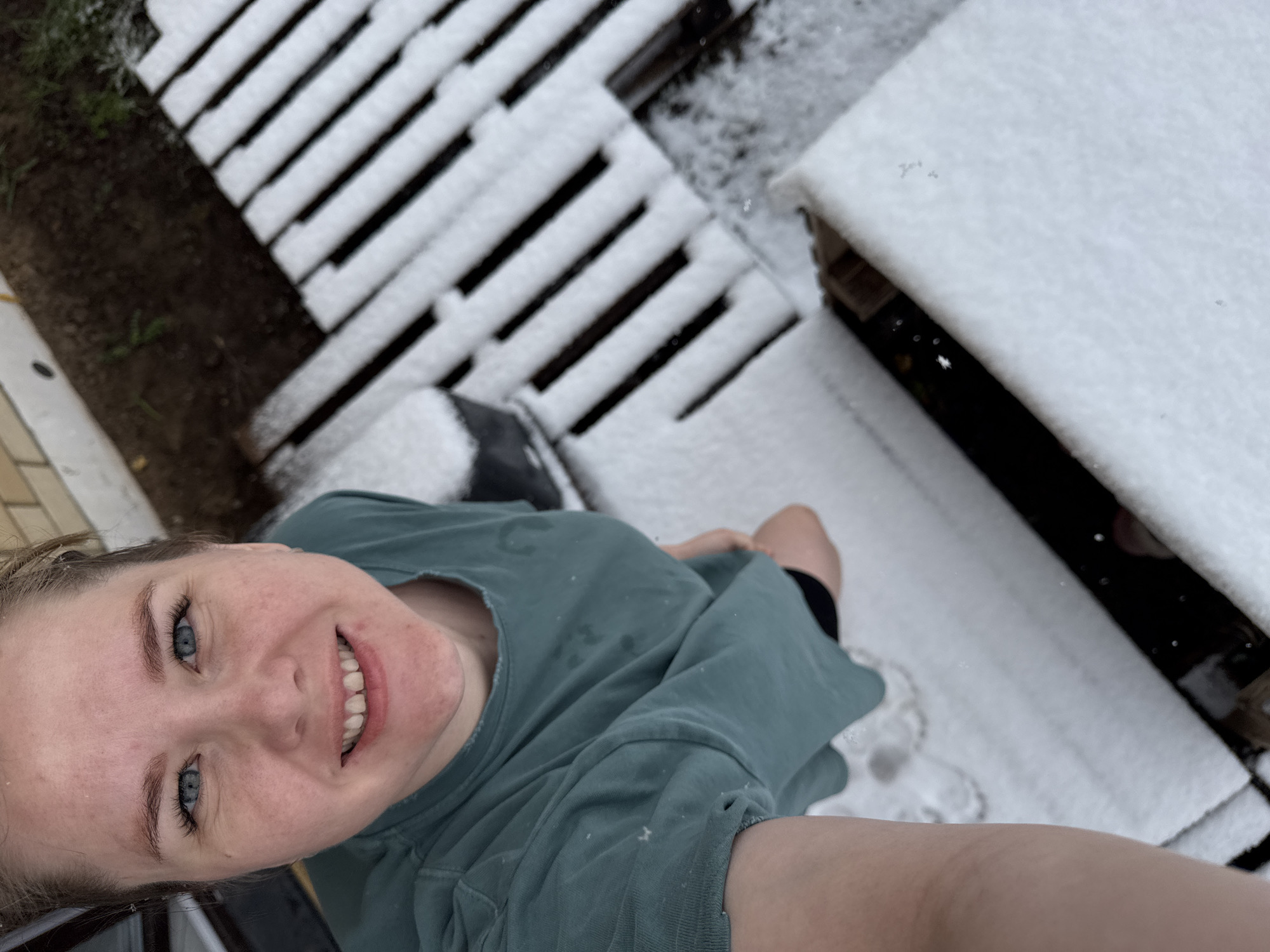 A young woman is taking a selfie outdoors on a snowy day. She is wearing a teal-colored t-shirt. Snow is visible on the ground and on a wooden structure in the background. The location is Gornyi (Dvurechenskoe S/P), Russia.