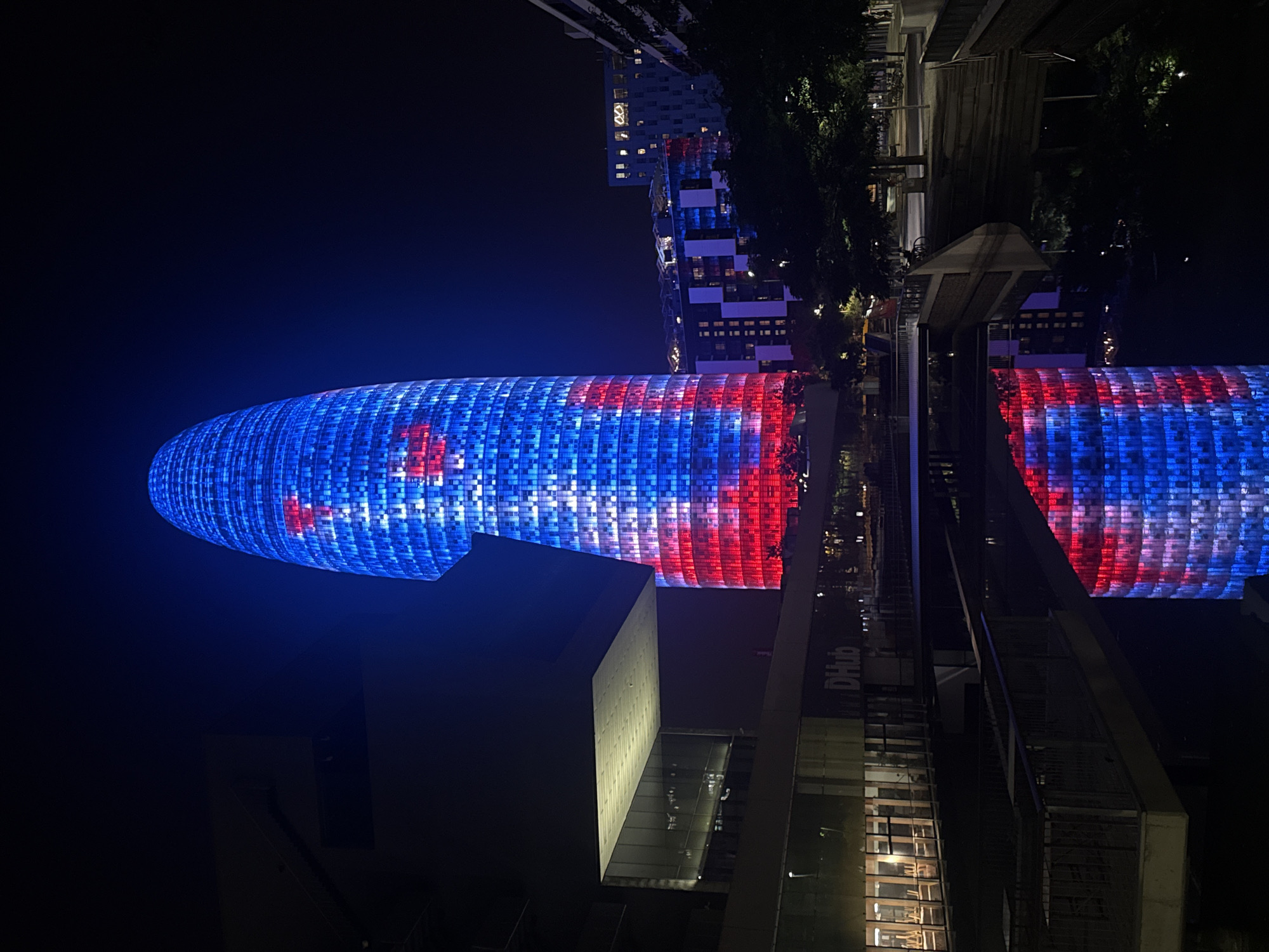 The image captures a nighttime view of the Torre Glòries, a skyscraper located in Barcelona, Spain. The building is illuminated with a striking pattern of blue and red lights, creating a vibrant display against the dark sky. The skyscraper's distinctive shape is visible, with its facade covered in a grid of illuminated windows that are highlighted by the colored lights.
In the foreground, a body of water reflects the colorful illumination of the skyscraper, creating a mirrored image that doubles the visual impact of the scene. The reflections are detailed and sharp, showing the architectural lines of the building and the patterns of light.
To the left of the skyscraper, a modern building with a cantilevered section is visible, also partially lit. This building features glass facades and a bright, illuminated ceiling or upper level. The text "DHub" is visible on this building.
Further in the background, other buildings and trees are present. Some buildings have their windows lit, adding to the urban nightscape. Trees with dense foliage are scattered throughout the scene, some silhouetted against the lights. A bridge or walkway crosses over the water, and on its far side, a street or path is visible with trees and what appear to be parked bicycles.
The overall impression is one of a dynamic and modern city at night, with the illuminated skyscraper serving as the central focal point. The scene is characterized by the interplay of light and shadow, architectural forms, and reflections in the water. There are no clearly discernible people in the image, though the presence of city infrastructure and potentially parked vehicles suggests urban activity.
