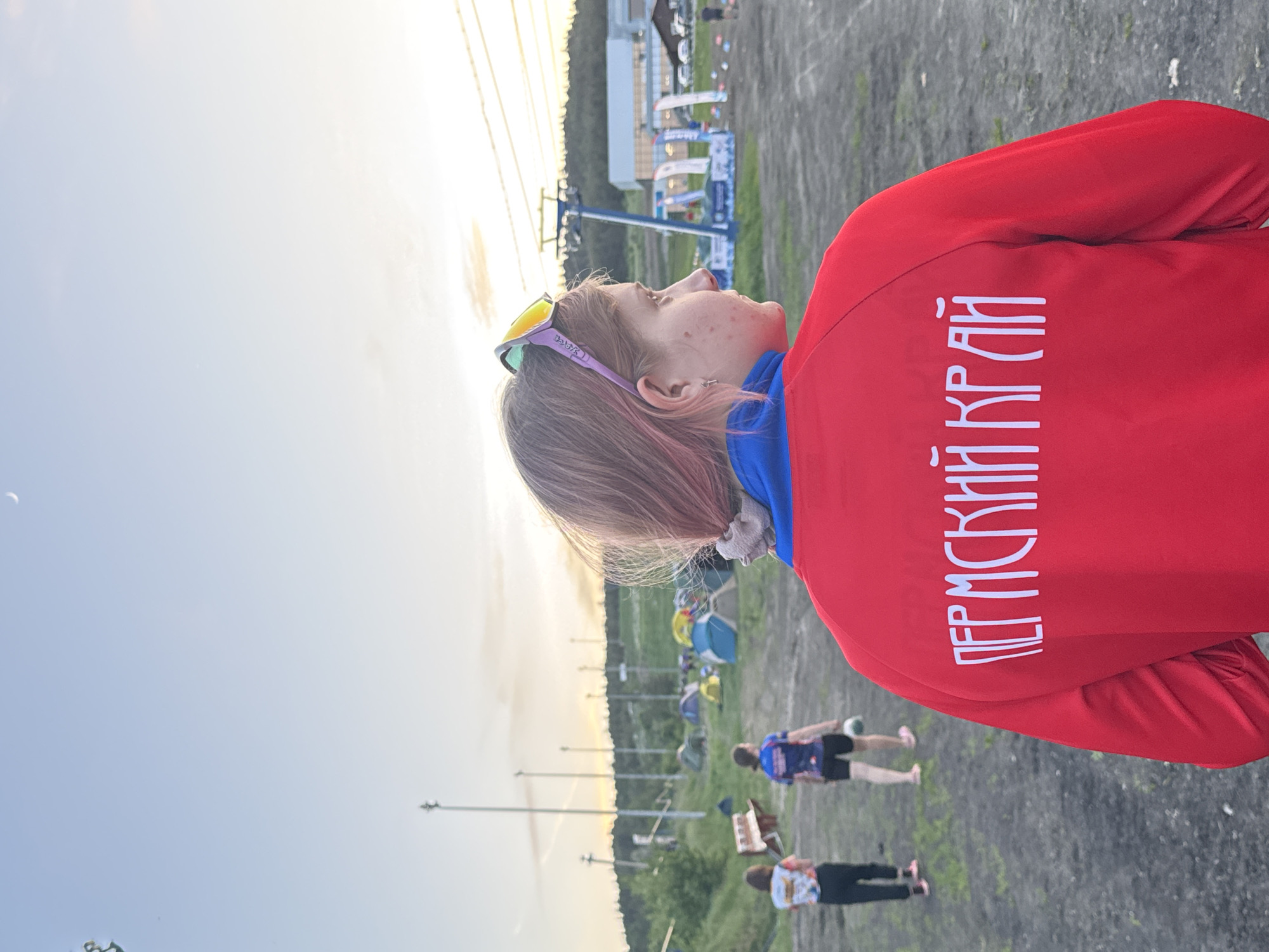 The image shows a young woman wearing a red jacket with the inscription "PERMSKIY KRAY" in white lettering. She has short, pink-tinted hair and sunglasses perched on her head. She is looking upwards towards a ski lift. In the background are tents, people, and a partially visible white building with flags. The scene appears to be outdoors, possibly at a sports event. Based on the jacket inscription and the presence of a ski lift, the location is likely near a ski resort within the Permskiy Kray region. The city of Khvalynsk is not evident in the image. The angle is low to the ground.