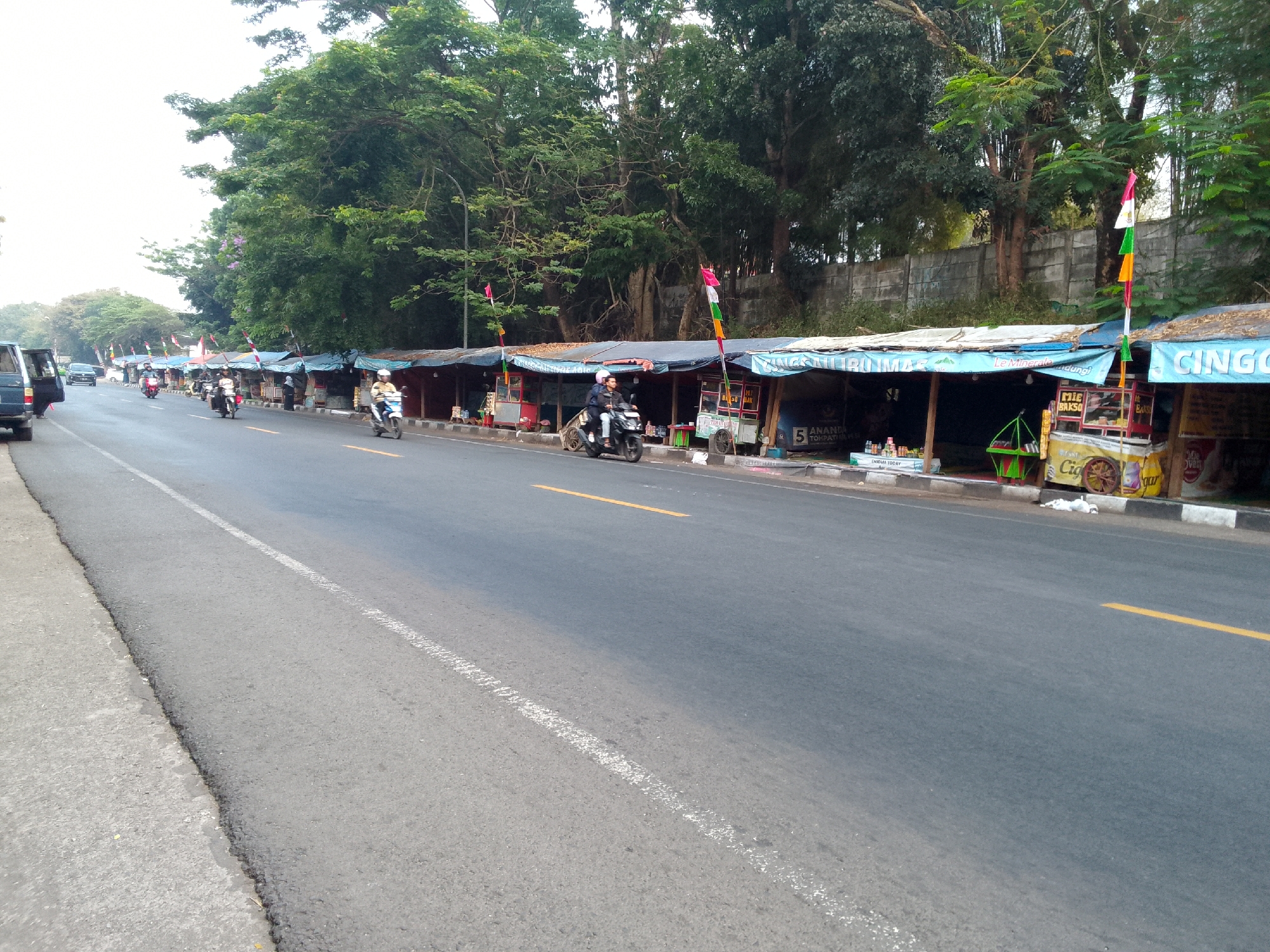 This is a wide-angle, eye-level shot of a road in Haurwangi, Indonesia, with a row of roadside food stalls lining the right side. The road surface is asphalt with white and yellow lane markings. On the left, a portion of a dark-colored van is visible, its driver's side door open. Several motorcycles are traveling on the road, some with riders. The stalls are covered with blue tarps, and some display flags. There are trees and a concrete wall in the background. The stalls appear to be selling food and drinks.