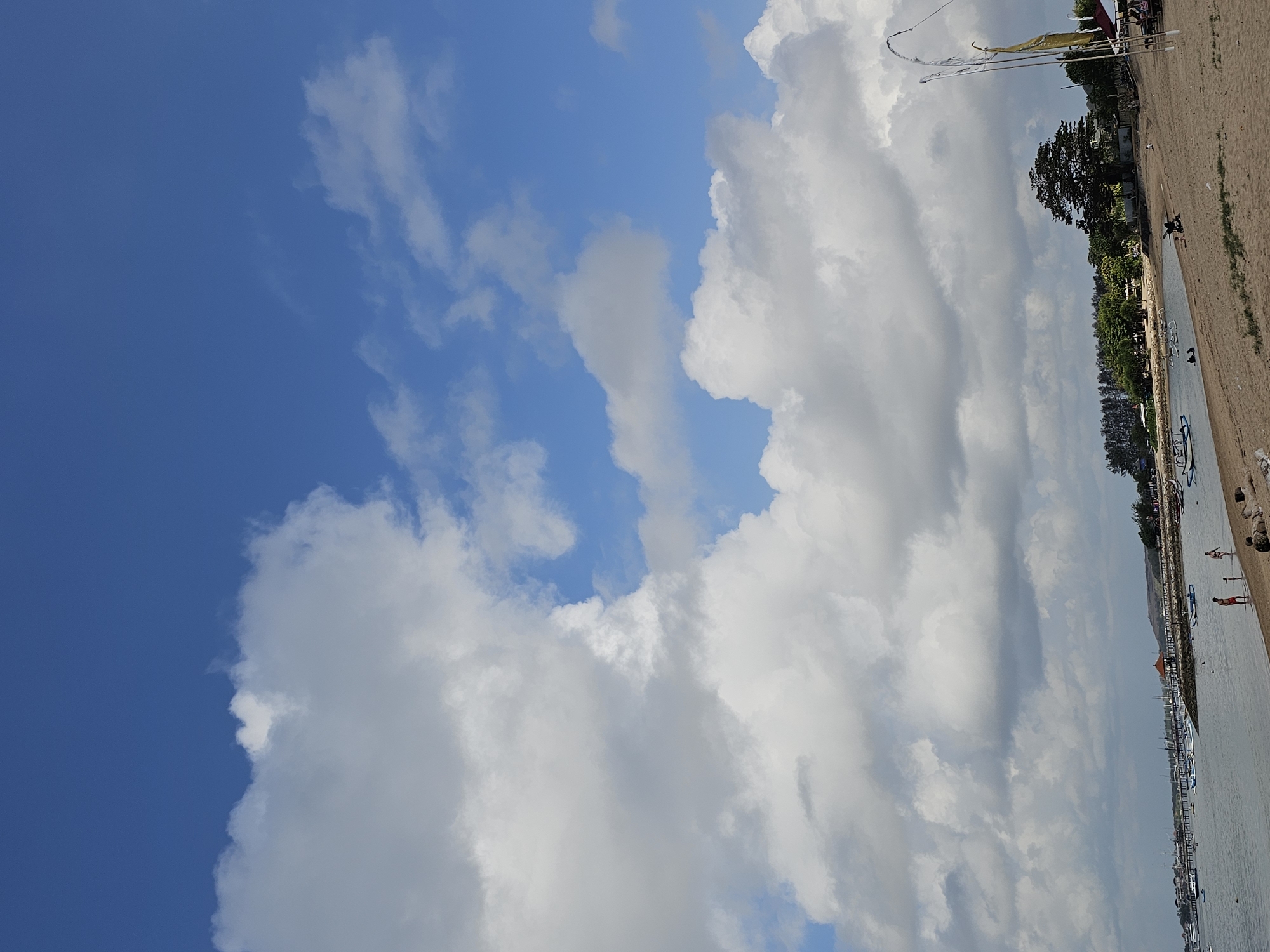 This image captures a scenic view of a beach in Denpasar, Indonesia, likely Sanur Beach, characterized by its calm waters and sandy shore. The sky is a prominent feature, filled with large, fluffy white clouds against a vibrant blue backdrop, suggesting fair weather.
In the foreground, a sandy beach stretches out, with some scattered logs and rocks. A few people are visible on the beach, including a child and two adults wading in the shallow water, and a person pushing a stroller further up on the sand.
The water is a tranquil blue, dotted with numerous small boats and a longer pier extending into the sea, lined with more vessels. A small gazebo-like structure sits near the water's edge on the right side of the image, with a few people seated under it.
The background reveals a lush line of trees, indicating a coastal vegetation zone, with some buildings nestled amongst them. The overall atmosphere is one of a relaxed day at the beach, with clear skies and calm conditions. The presence of traditional Indonesian boats and the tropical greenery strongly suggest the location is Denpasar, Indonesia.