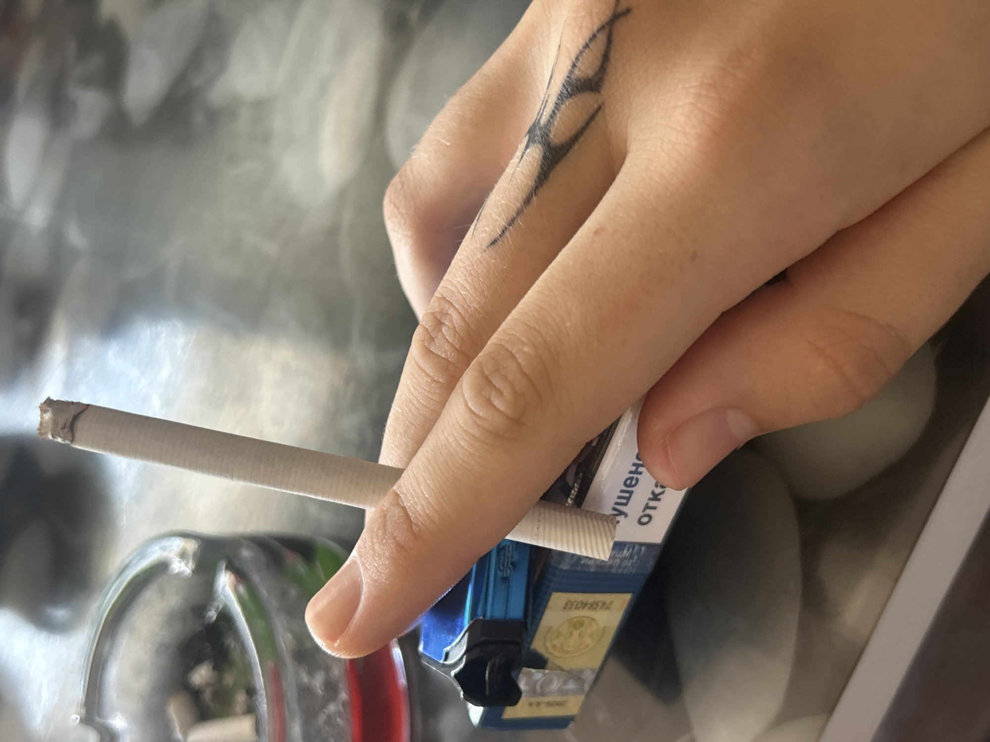 The media file captures a close-up view of a person's hand holding a lit cigarette and a blue cigarette pack. A prominent black, tribal-style tattoo adorns the back of the hand, extending across the knuckles. The cigarette, with a small amount of ash at its tip, is held between the fingers.
The blue cigarette pack features visible text in Cyrillic script, including partial words such as "УШЕН" and "ОТКАЗ," which are likely components of a public health warning. A golden emblem or seal is also discernible on the pack.
In the background, a clear glass ashtray with some residual ash and cigarette butts is partially visible. The surface beneath the hand and objects appears reflective, showing blurry, indistinct shapes that suggest other items, possibly eggs, as indicated by a provided crop. This indoor scene, characterized by the Cyrillic text on the cigarette packaging and associated with the location context of Loznitsa, Bulgaria, points to a setting in a region where Cyrillic script is used. The image depicts a casual moment related to smoking. No specific time of day or emotional state is discernible.