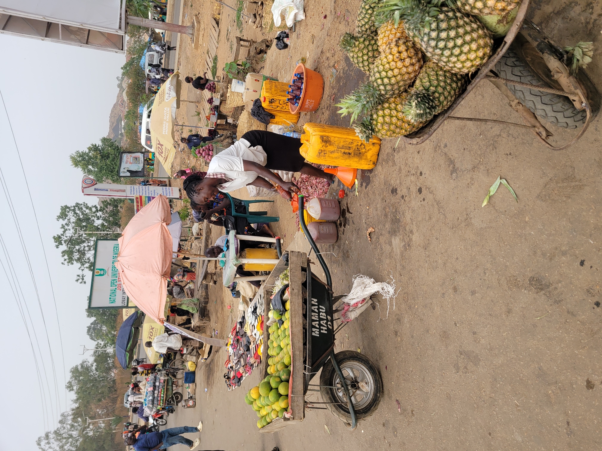 The image captures a vibrant street market scene in Jos, Nigeria, during daytime under clear skies. The setting is a bustling roadside with numerous vendors and shoppers.
In the foreground, a woman in a white top and dark skirt is engaged in an activity on the ground, possibly sorting or processing items, with several yellow jerry cans, funnels, and an orange tub labeled "HAPPY" filled with bottles nearby. To her left, a wooden cart prominently displays "MAMAN HABU 01" and is laden with green and yellow citrus fruits, likely oranges or limes. On the right, a different cart or wheelbarrow is overflowing with ripe pineapples. Red onions are visible in a container near the woman.
Throughout the scene, several people are observed, some sitting under various umbrellas (pink, blue, and a beige one with "10X MORE DATA MORE VALUE" and "Glo Cake Amm" branding), suggesting different stalls. Other individuals are standing or moving between stalls, engaging in commerce.
In the background, road signs are visible, including one for "NATIONAL OPEN UNIVERSITY OF NIGERIA JOS ST LOMAY INT" and another for "Bimme Hospital," confirming the urban context. A partial view of a white van and motorbikes indicates vehicular traffic on the road. The overall atmosphere suggests a typical day of trade and daily life in the market.