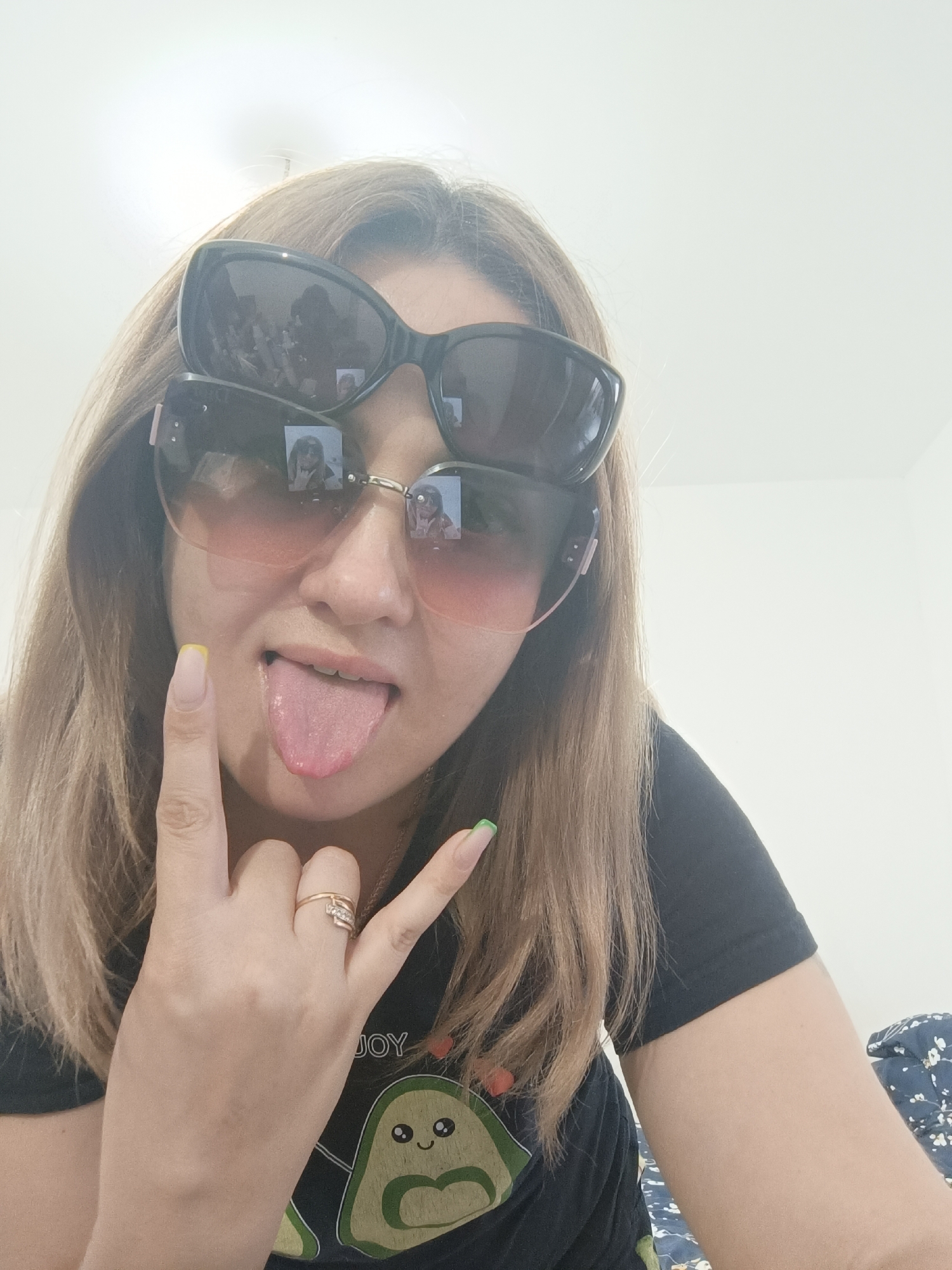 A woman with shoulder-length brown hair is pictured from the chest up, wearing two pairs of sunglasses, one stacked on top of the other. She has her tongue sticking out and is making a rock-and-roll hand gesture with her right hand, while her left hand is raised with her index finger pointed upwards. Her nails are painted, with yellow tips on her left hand and green tips on her right. She is wearing a black t-shirt with a graphic print of an avocado. The background appears to be an indoor setting with a plain white wall. There is no identifiable information suggesting the location is Taraz city administration, Kazakhstan.