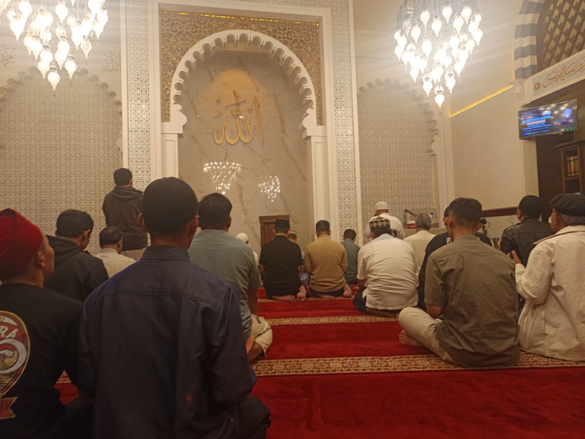 A group of Muslim men are gathered inside a mosque, praying. The men are kneeling on a red carpet with intricate patterns. The mosque's interior features ornate white and gold arches, and a large chandelier hangs from the ceiling. In the background, there is a television screen displaying what appears to be a religious program. The scene suggests a prayer service is taking place in a well-decorated mosque.