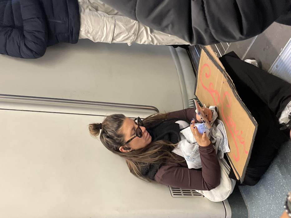 The image displays the interior of a subway car in Barcelona, Spain. A woman with long brown hair, secured in a half-up top knot, sits on the blue-speckled floor, leaning against a light gray wall. She is wearing dark oval sunglasses, wireless earbuds in both ears, a dark brown long-sleeved top, a black scarf, black pants, and white sneakers. A light blue denim jacket is draped across her lap, partially covering a light-colored shoulder bag positioned next to her left hip. Her fingernails are painted purple as she holds and intently interacts with a white smartphone with both hands. In front of her, resting on her lap and the floor, is a rectangular piece of brown cardboard with a pink/red stylized design or script and black-painted edges. To her immediate right, a standing individual is partially visible from the mid-torso down, wearing a dark blue puffer jacket and light-colored cargo pants. The lower portion of another person in dark clothing is visible in the bottom right foreground. A white ventilation grate is present on the light gray wall to the woman's left. A vertical metal support or door frame runs along the wall to her right.