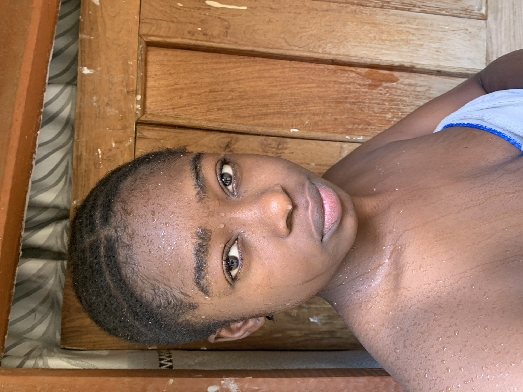 A close-up, high-angle shot depicts a young Black person with short, styled hair, lying on a wooden surface. The person is partially covered with a white towel with blue trim. Droplets of water are visible on their face, head, and chest. The background features a wooden frame and striped material, suggesting a bed or seating area. The image was taken indoors, location unknown. The city of Rafin Par, Nigeria, is not visible in the image. There are no actions, interactions, or other people in the image.