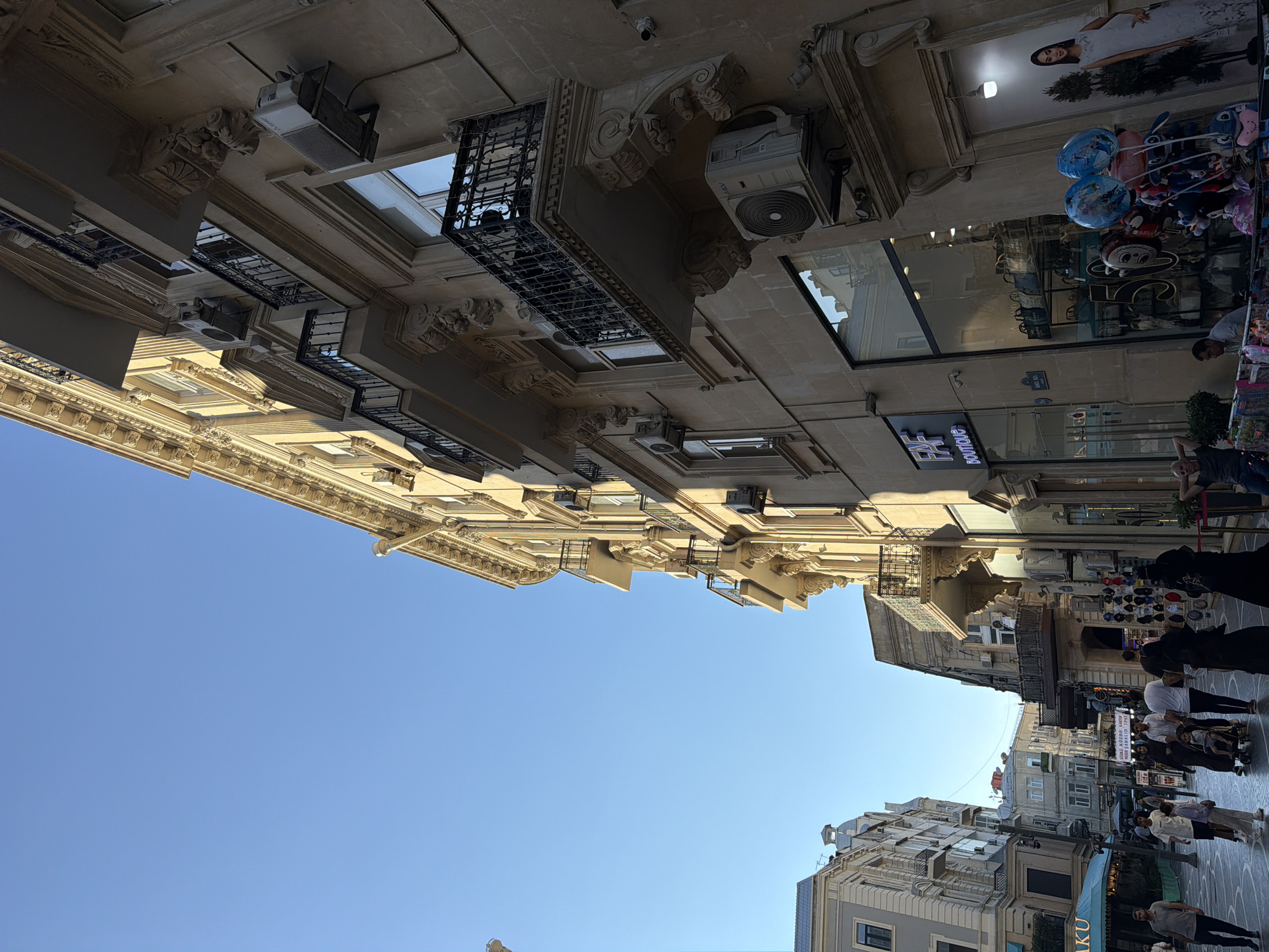 In the city of Sabail Raion, Azerbaijan, on a clear day, a low-angle shot looks up at a grand, beige stone building with ornate architectural details and multiple balconies. The building is a prominent feature of a bustling street, evidenced by the shadows cast by its upper levels and the presence of people walking below. The street level of the building features storefronts, including one with a sign reading "FAF BOUTIQUE" and a large display window showcasing handbags. Adjacent to the boutique, another storefront window displays a mannequin in a white dress. Further down the street, other buildings are visible, suggesting a dense urban environment. Several people are seen walking on the street, with some carrying shopping bags. The overall scene depicts a lively urban setting with a mix of old-world architecture and modern commercial activity.