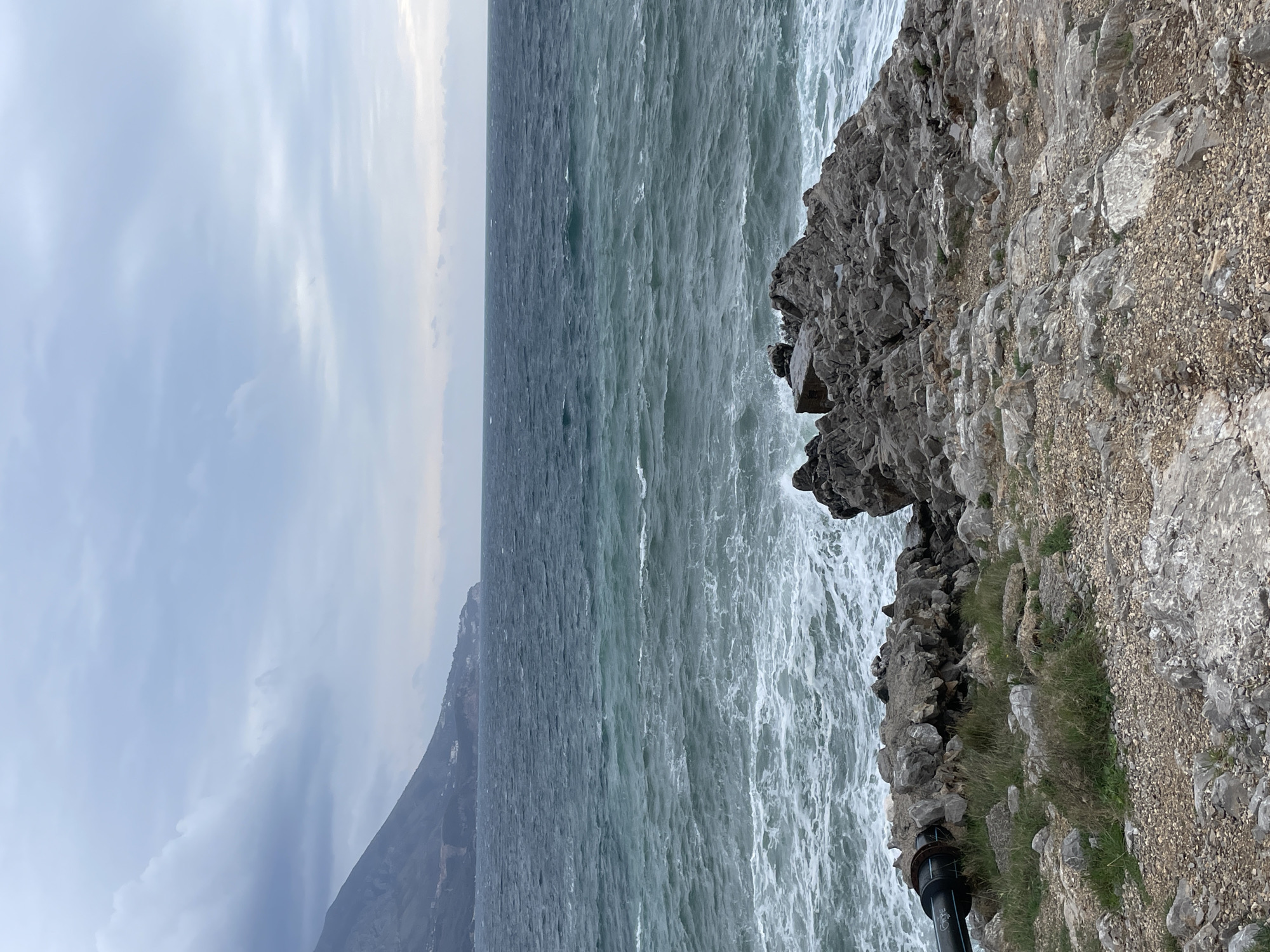 The image depicts a turbulent coastal scene in Budva, Montenegro. The foreground shows a rocky, uneven shoreline with sparse patches of green vegetation and gravel. A dark-colored pipeline, possibly an outfall, is partially visible protruding from the left side and extending towards the water's edge, partially obscured by rocks. The midground features a rough, dark teal-colored sea with numerous whitecaps and breaking waves actively crashing against the rocks. The background reveals a cloudy, overcast sky, predominantly light grey. In the distant left, a mountainous coastline is visible, featuring scattered buildings on its lower slopes. No individuals or vessels are present in the observable scene.