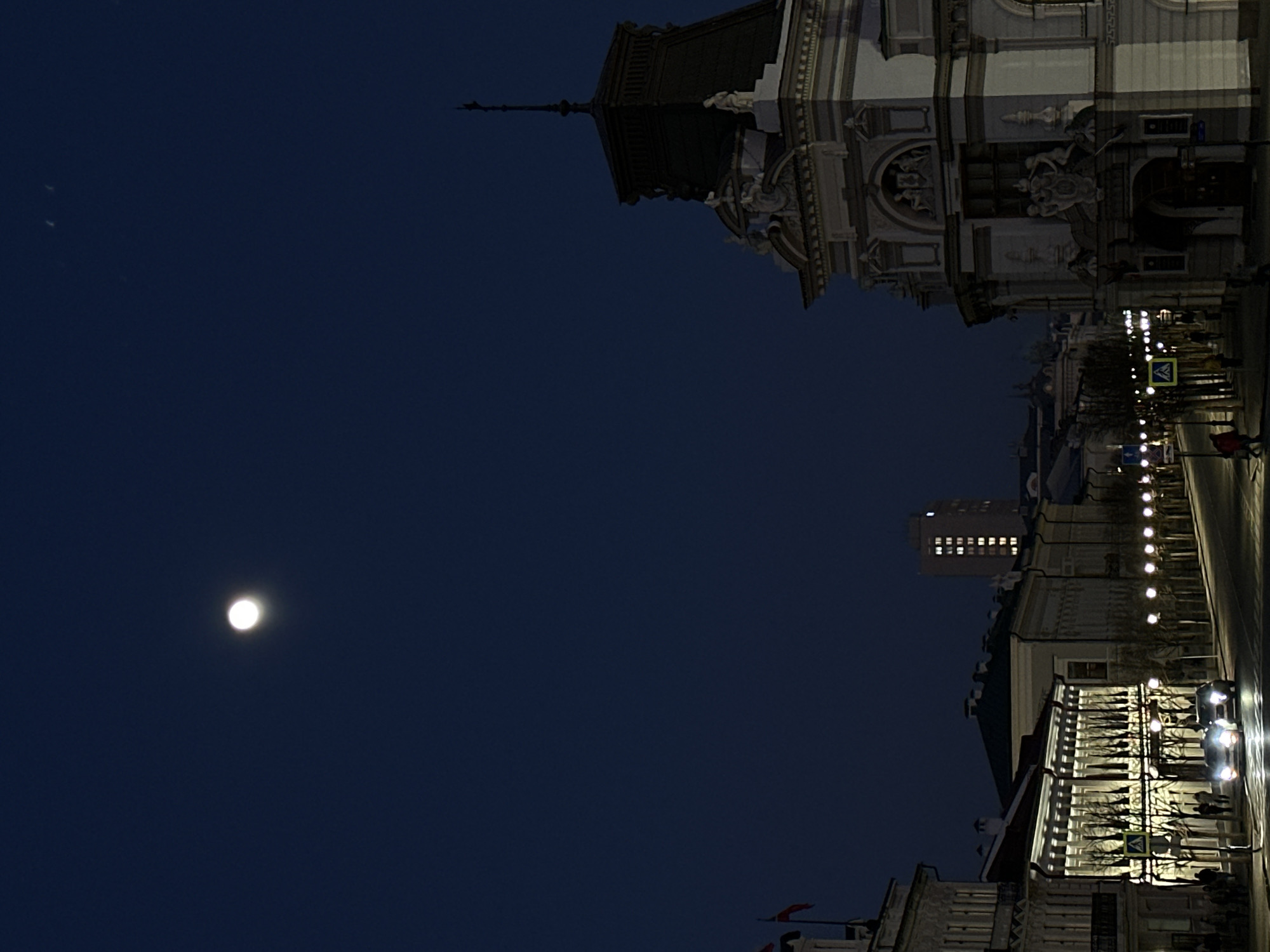 At night in Kazan, Russia, the moon is visible in the night sky. The scene showcases illuminated architecture, streetlights along a road, and several individuals walking in the distance. A pedestrian crosswalk is indicated by a green and yellow sign. The architecture features ornamental details and intricate designs. A taller building is also visible.