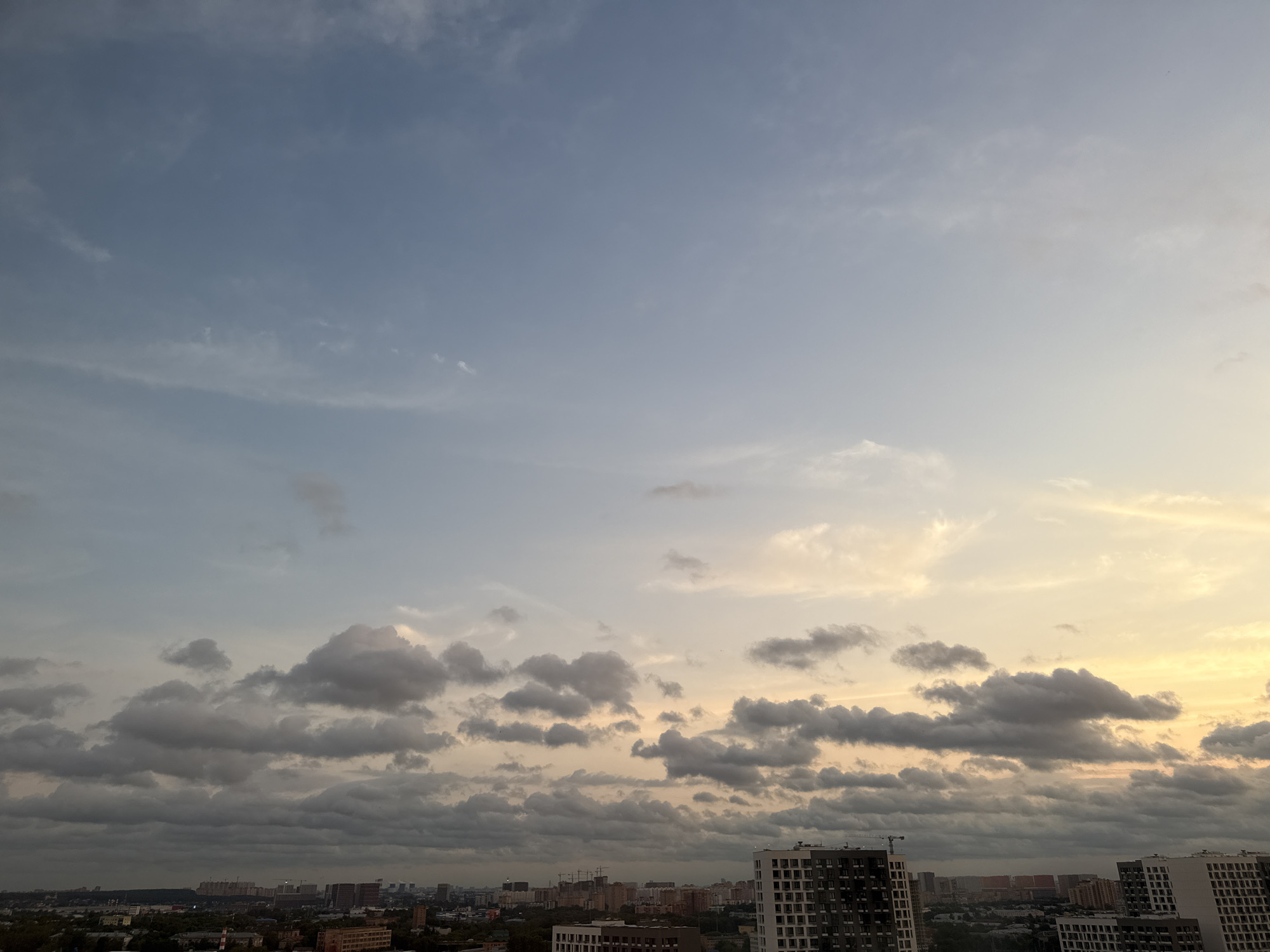 This is a medium-shot, high-angle view of a cityscape in Lyubertsy, Russia, under a sky filled with clouds. The sky transitions from a clear, pale blue at the top to hues of yellow and orange near the horizon, suggesting either dawn or dusk. Wispy white clouds are visible in the upper portion, while heavier, gray clouds dominate the middle and lower parts of the sky, obscuring the sun.
The city below consists of a dense collection of buildings of various heights and architectural styles. Modern high-rise apartment buildings with visible windows and balconies are prominent, particularly in the foreground and right side of the frame. Taller structures, possibly under construction or industrial facilities with cranes, are discernible in the mid-ground. Lower buildings and trees are scattered throughout the background. The overall atmosphere is muted and overcast due to the cloud cover. There are no visible people or distinct actions occurring within the frame. The focus is on the interplay between the urban landscape and the dynamic sky.