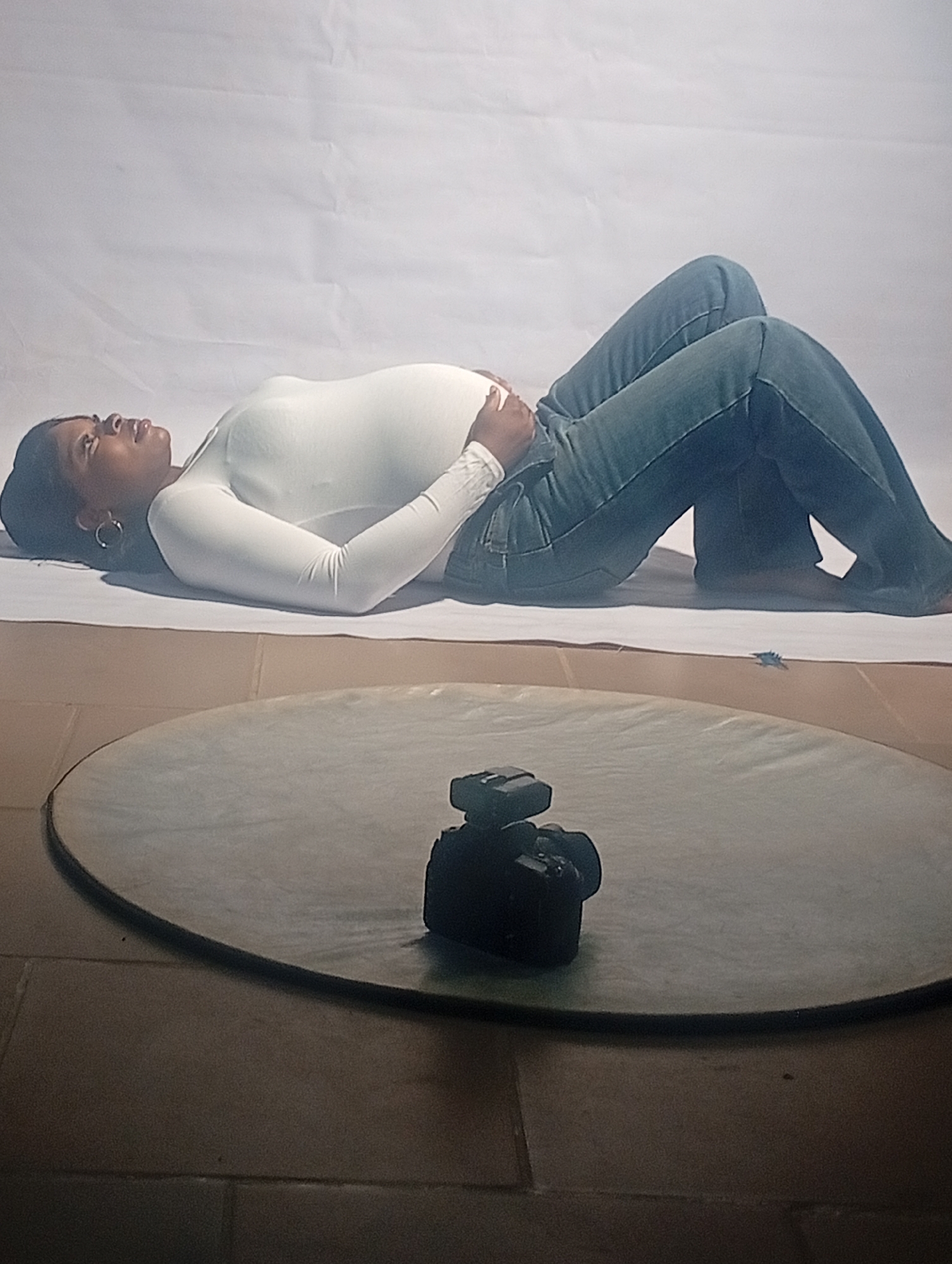A pregnant woman lies on a white sheet on the ground. She is wearing a white long-sleeved shirt and blue jeans. Her hands are resting on her stomach. A camera is positioned on a circular reflector. The scene appears to be indoors, with a tiled floor. The location is Barakin Akawo, Nigeria.