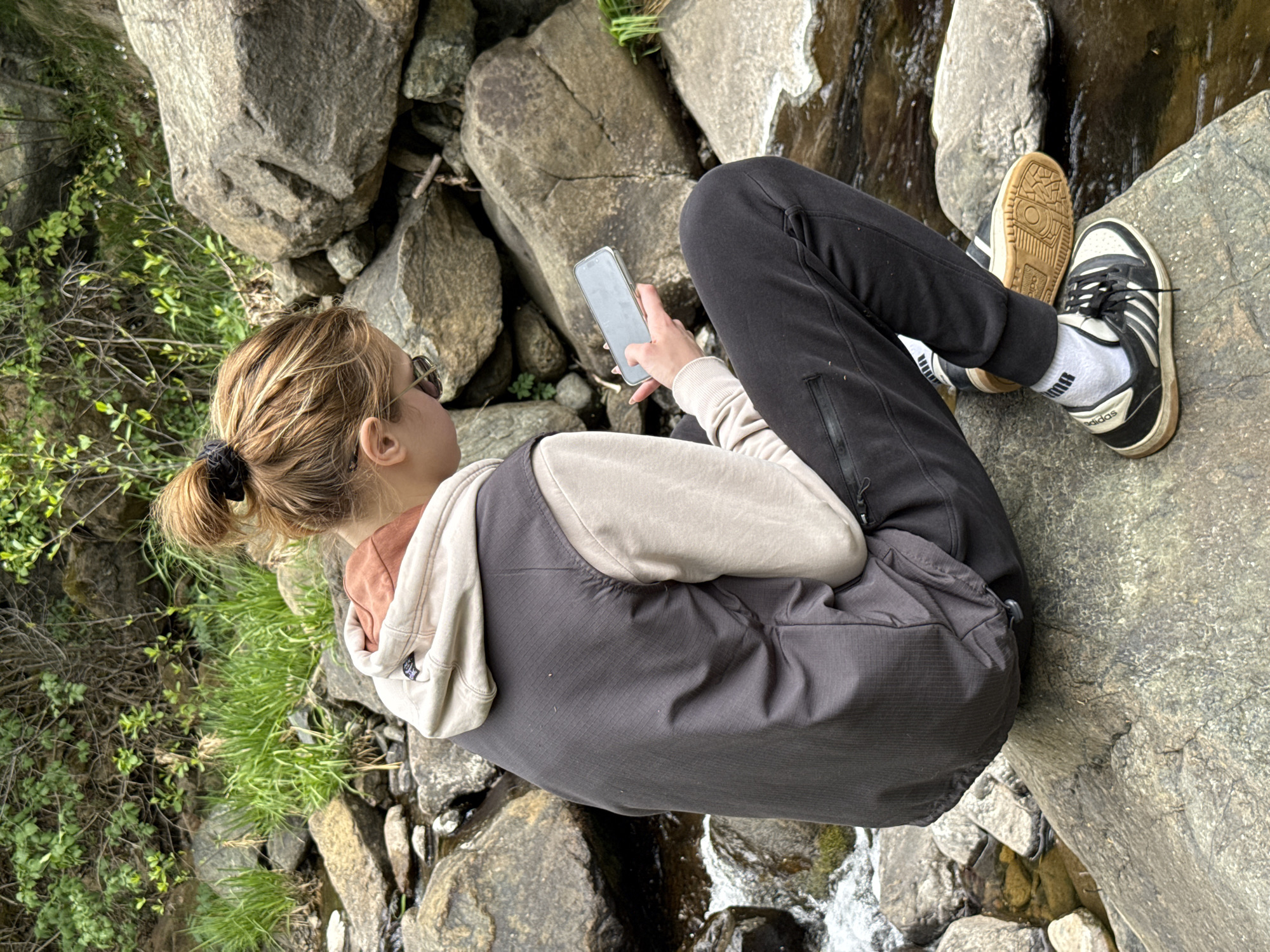 The image shows a person sitting on rocks next to a small stream, possibly near Maglič, Serbia, given the surrounding mountainous terrain and rock formations. The person is wearing black athletic pants, a light-colored long-sleeved shirt under a dark-colored vest with a hood, and black and white Adidas sneakers. They have fair skin and blonde hair pulled back in a ponytail. The person is looking at a mobile phone held in their hands. The environment includes large rocks, green vegetation, and a stream with flowing water.