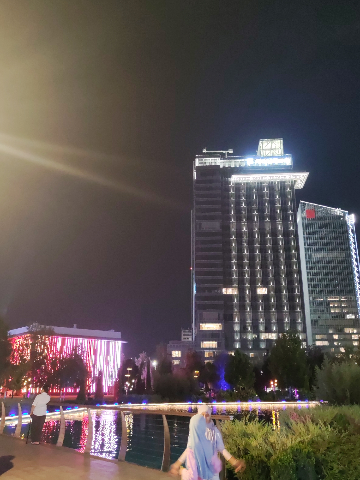 The image shows a nighttime scene in the city of Tashkent, Uzbekistan, likely in a modern, well-lit urban park or plaza area. Dominating the background are two tall, modern buildings. The building on the left is a high-rise with a dark facade, illuminated by numerous vertical rows of bright white lights. A second, slightly shorter building stands to its right, also featuring a glass facade and illuminated windows.
In the foreground, a body of water, possibly a pond or canal, reflects the lights from the surrounding environment. Along the edge of the water is a paved walkway where two individuals are visible. On the left, a person wearing a white shirt and dark pants stands with a cane, looking towards the illuminated building on the left. Further to the right, a person wearing a light blue hijab and a blue hooded garment is partially visible, appearing to be walking or posing with their arms outstretched.
To the left of the water and in front of the first tall building, there is another, lower building or structure adorned with vertical LED lights in shades of pink and red, creating a vibrant, illuminated effect. Trees and greenery are scattered throughout the scene, softening the urban landscape. The overall atmosphere is one of a lively, illuminated public space at night.