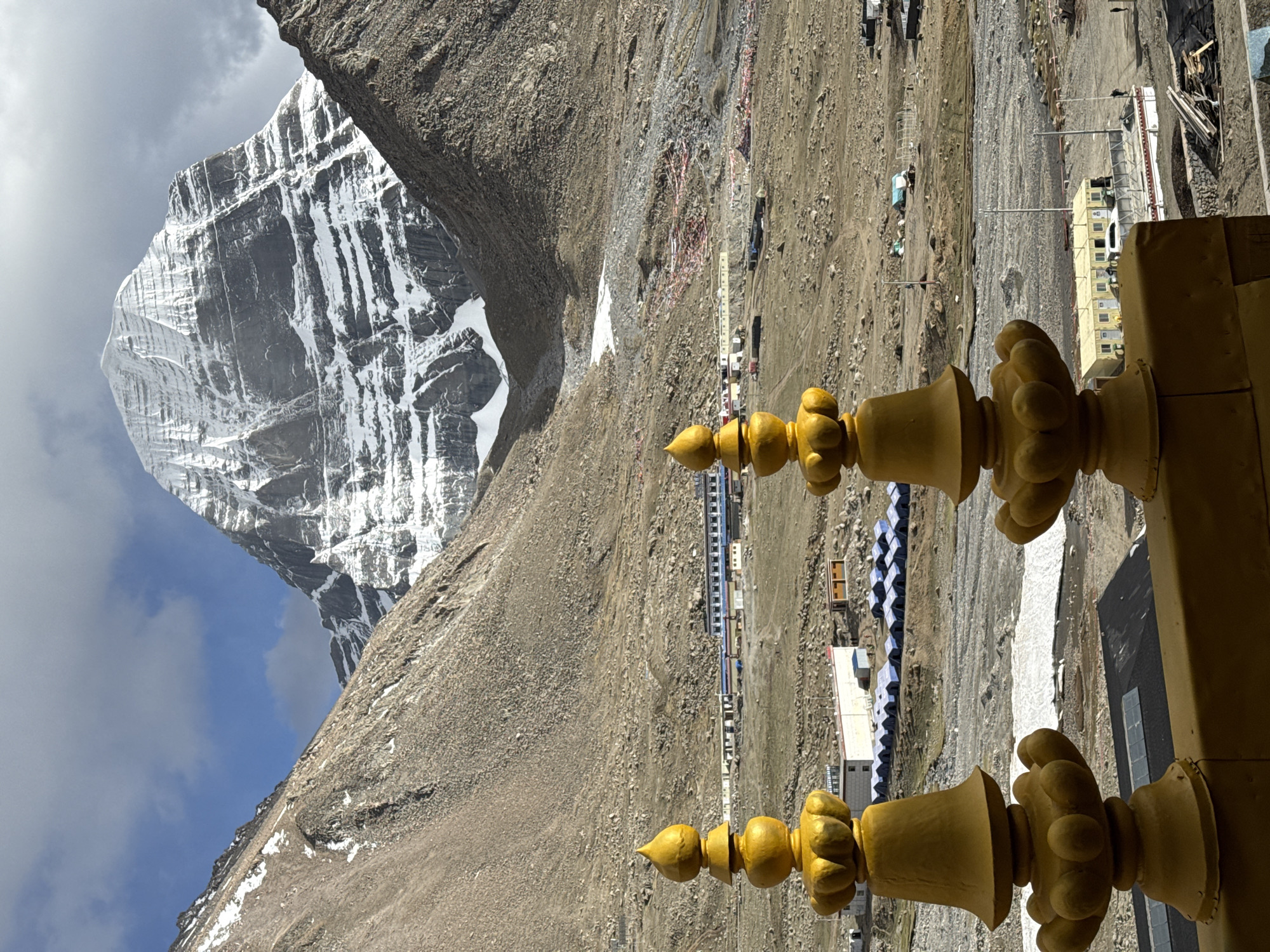 The image captures a mountainous landscape, likely in Tibet, framed by a gilded architectural element in the foreground. A prominent snow-capped mountain rises to the left, partially obscured by a steep, rocky slope. The lower section of the landscape reveals a settlement or base camp, with buildings and tent-like structures. Prayer flags are visible along the slope leading towards the mountain. The gilded element in the foreground appears to be part of a balustrade or roof ornament, suggesting the photograph was taken from an elevated vantage point. The sky is partly cloudy.
