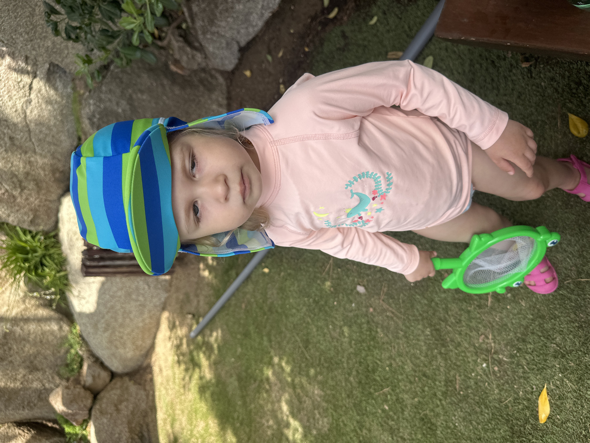 A young child wearing a pink long-sleeved rashguard with a whale design and a blue and green striped sun hat stands on a patch of artificial turf. The child holds a green turtle-shaped net in their right hand and is wearing pink sandals. In the background, large rocks and some greenery are visible, suggesting an outdoor setting, possibly a garden or a playground area. A gray pipe or rod runs across the ground behind the child. The lighting indicates it is daytime, with shadows cast on the grass. The child's expression is neutral. The scene appears to be located in Sant Feliu De Guíxols, Spain, given the context.
