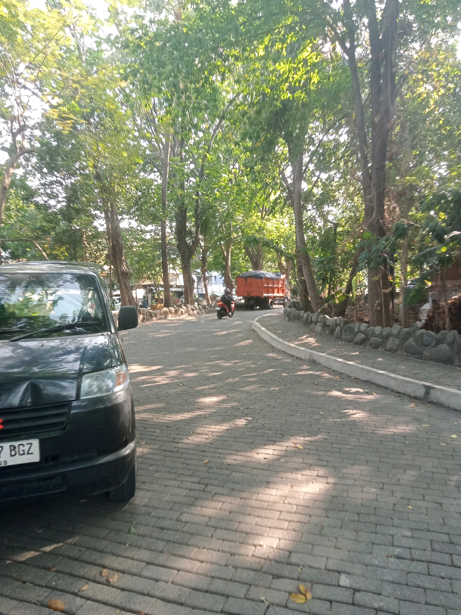 The image shows a paved road in North Jakarta, Indonesia, lined with trees. A dark-colored van is parked on the left side of the road, with its front visible. Further down the road, an orange truck is driving away from the camera, and a person on a motorcycle is also visible. The road surface is made of interlocking bricks, and there are shadows cast by the trees on the ground. The area appears to be a park or a residential area with a lot of greenery.