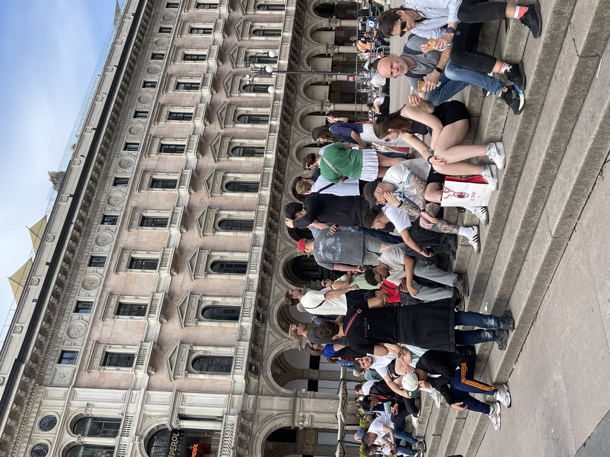 The image depicts a crowd of people sitting on steps in Milan, Italy. They are positioned in front of a tall, ornate building with multiple arched windows and detailed facade work. Many people are facing forward, observing the surroundings, while some are conversing with each other. The steps they are sitting on appear to be concrete or stone. The scene is outdoors, with daylight visible in the sky.