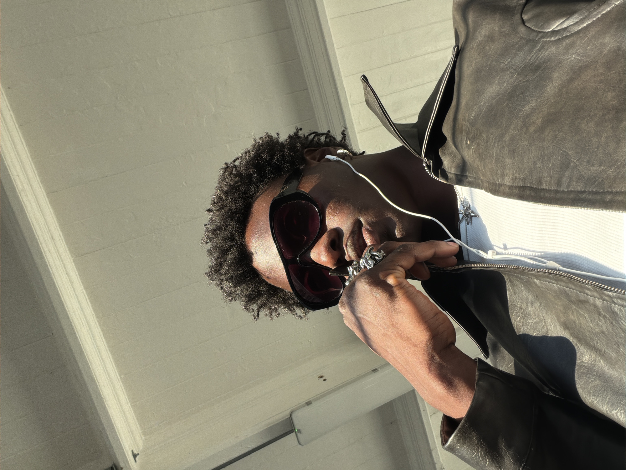 A low-angle, sunlit portrait captures a Black man from the chest up, positioned against a light-colored, planked overhead structure. He has short, curly dark hair and wears large, dark-framed sunglasses that cover a significant portion of his face. White wired earphones are visible in his ears, with the cord extending downwards against his skin and the white ribbed shirt he wears under an unzipped dark grey or black leather jacket. His right hand, adorned with several silver-toned rings, is raised near his face, with fingers appearing to adjust the earphones or sunglasses. The bright sunlight creates strong highlights on his skin and hair and casts distinct shadows. The background shows horizontal wooden planks or siding overhead, with a white rectangular device, possibly a sensor or light fixture, featuring a green indicator light, affixed to its underside. The image is taken in clear, bright weather. No text is visible in the frame.