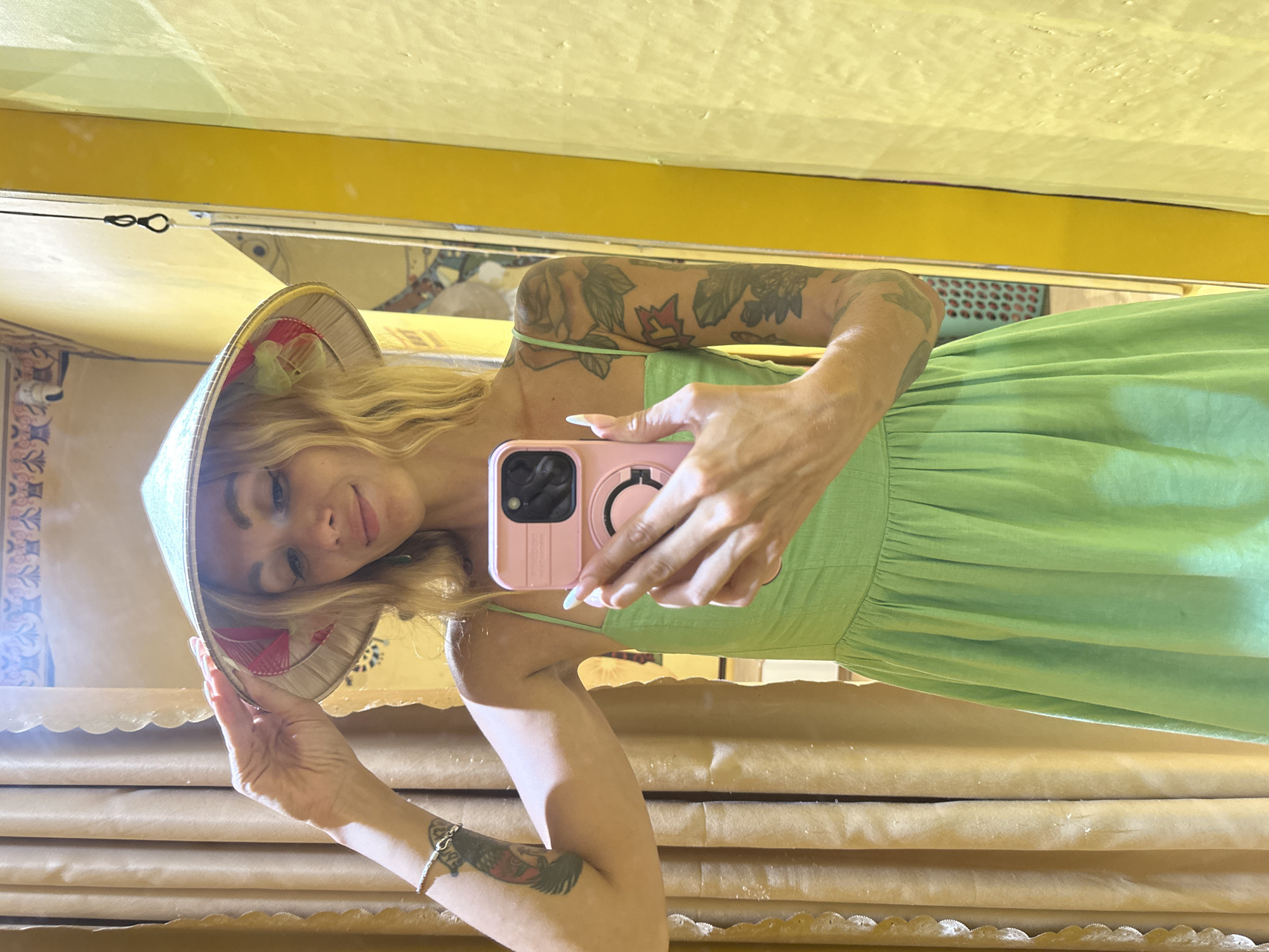 In an indoor setting, possibly a fitting room, a woman with blonde hair and visible tattoos on her arms is taking a mirror selfie. She is wearing a light green sundress and a conical hat. She holds a pink smartphone in her right hand, angled to capture her reflection. The background features textured tan curtains to her left and a yellow wall to her right. The overall lighting is warm and slightly diffused. The location is identified as Phu Thuy Ward, Vietnam.
