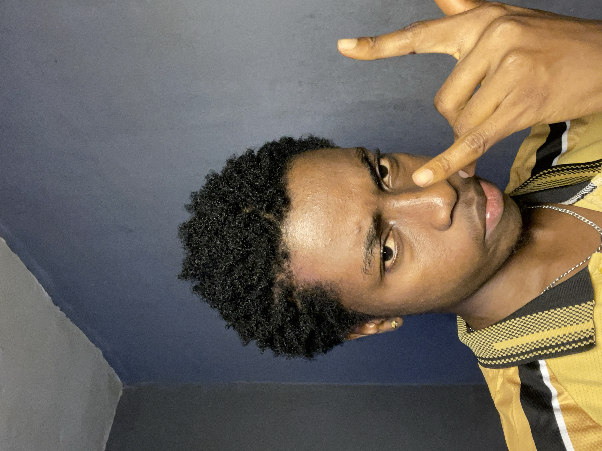 A medium shot shows a young man posing for the camera. His short, black hair is styled in tight coils. He has dark skin and brown eyes. He's wearing a yellow and black shirt. His index finger touches his nose, while another finger points upwards. A silver chain adorns his neck. The background is a dark blue and gray wall. The image does not provide information about the city of Nkpor, Nigeria.