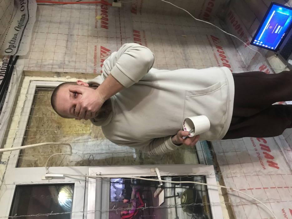 The image captures a man standing indoors, in what appears to be a room under renovation or construction. The man is positioned centrally, facing slightly towards the left of the frame with his head turned towards the viewer. He has a shaved or very short haircut and a light beard or stubble. He is wearing a light-colored, possibly off-white or beige, hooded sweatshirt and dark trousers. His right hand is raised, covering his mouth and part of his nose, an expression that might convey surprise, disbelief, or an attempt to stifle a laugh or comment. In his left hand, he holds a white mug with a spoon inside.
The setting reveals walls covered with a grid-patterned material, likely an insulating or vapor barrier membrane, consistent with building construction. Visible text on this material includes "ИЗОСПАН", "Isospan", "www.isospan.ru", and "сделано в России" (made in Russia), indicating Russian-made building materials. A white UPVC window or door frame is visible in the lower left portion of the image, adorned with thin string lights or wires. Through the glass, reflections and hints of an interior space can be seen. Various white and black electrical wires or cables are strung across the room, some hanging loosely, others appearing to be temporarily fixed to the walls.
In the upper right corner of the image, a laptop rests on a surface, with its screen illuminated and displaying a blue interface, possibly a login screen or a program. The lighting in the room appears to be artificial, suggesting an indoor environment without strong natural light, or possibly nighttime. The overall atmosphere points to a casual moment, perhaps a break during work in an unfinished or partially completed space.
