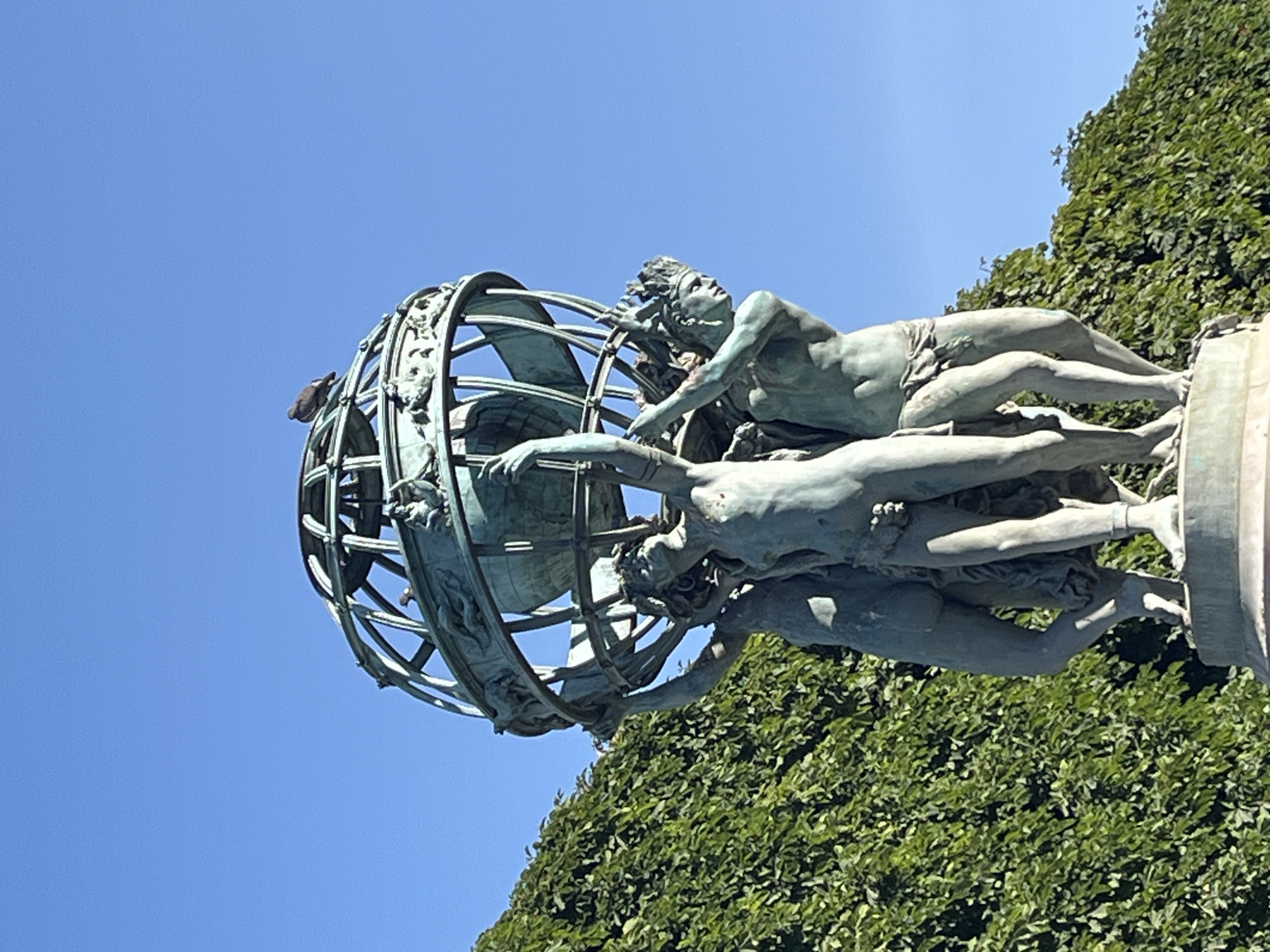 The image shows a bronze sculpture of three figures holding up a globe. A pigeon is perched on top of the globe. The sculpture is set against a backdrop of lush green foliage and a clear blue sky. This sculpture is located in the Luxembourg Gardens in Paris, France.
