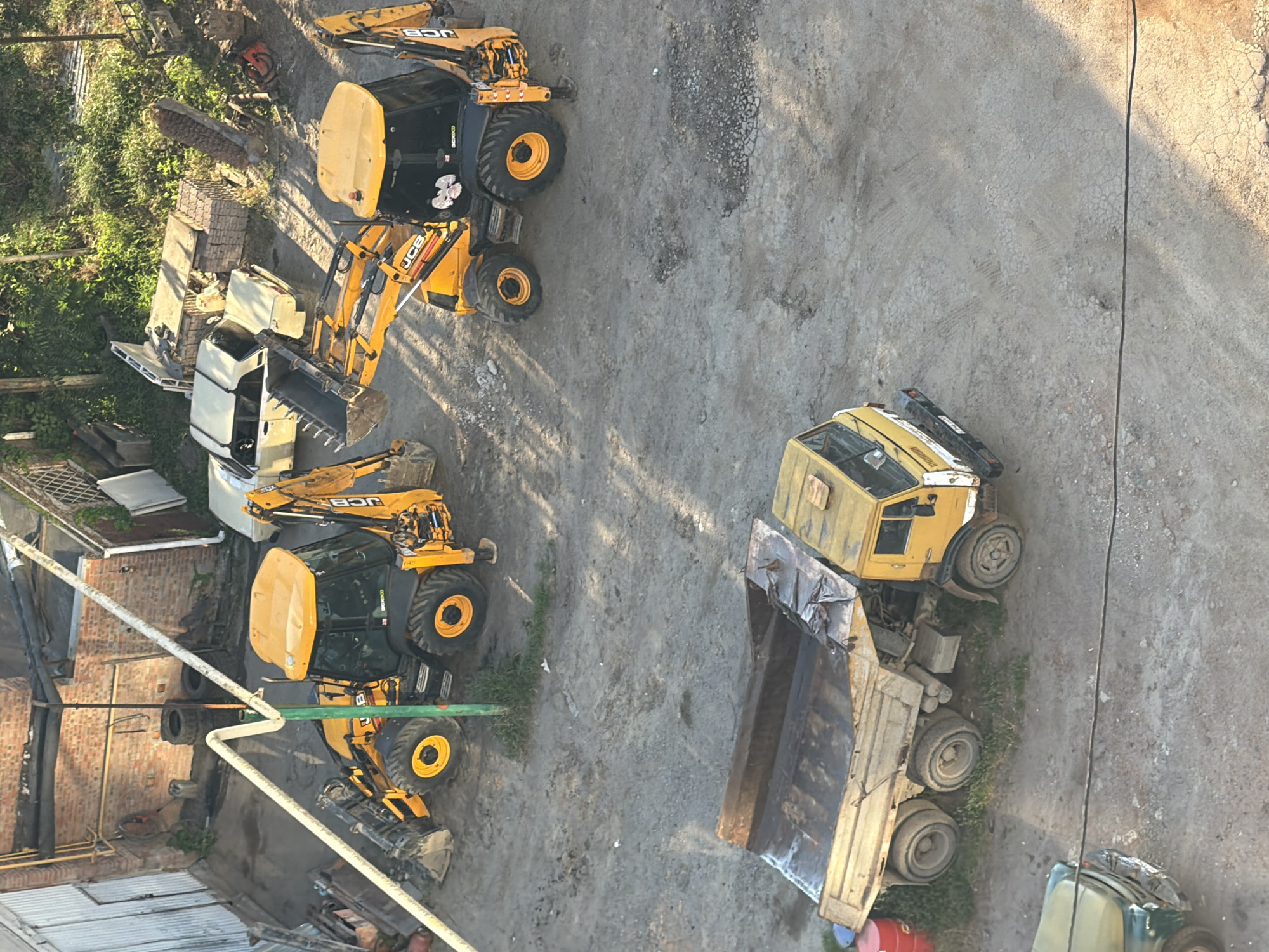 This media file shows a construction site with several yellow JCB backhoe loaders and a yellow dump truck parked on a dirt and gravel surface. The machines are positioned at various angles, suggesting they are either idle or have been recently moved. The scene is outdoors, with sunlight casting long shadows, indicating it is likely morning or late afternoon. Lush green foliage is visible on the left side of the image, suggesting the site is in a semi-rural or developed area. There are also some brick structures and industrial piping present, adding to the construction site context. The ground is uneven and appears to have tire tracks from heavy machinery. A small portion of a green vehicle and a red barrel are visible at the bottom right, adding to the variety of objects on the site. No people are visible in the image, and the overall impression is one of a quiet, possibly abandoned or off-duty, construction area. The visible text on the machines appears to be "JCB." The location context provided is Ovoshchnoy, Russia.