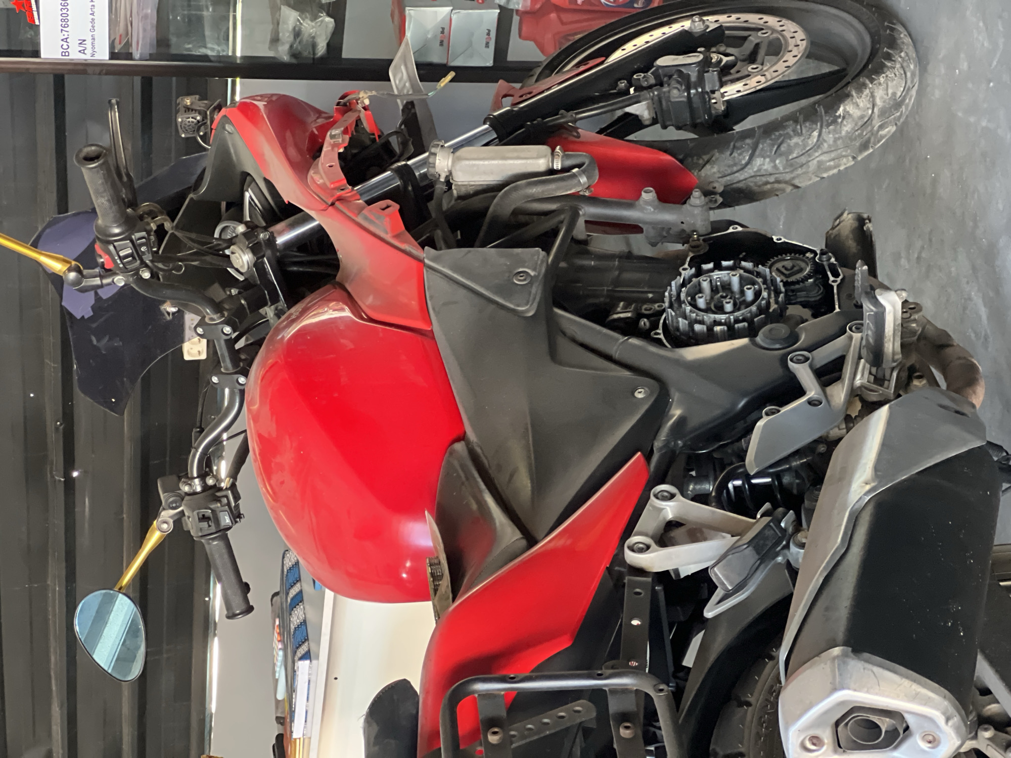 A red motorcycle is parked in what appears to be a garage or workshop. The motorcycle is the primary subject, angled towards the viewer's left. Its front fairing appears to be damaged or removed, revealing internal engine components. The motorcycle has a black seat, a black exhaust pipe, and a silver luggage rack on the left side. The background shows shelves with various items, including boxes and bottles. A sign with Indonesian text and numbers, possibly related to banking or a business, is visible on the right. The floor is a gray concrete surface. The scene suggests a motorcycle repair or storage environment in Denpasar, Indonesia.