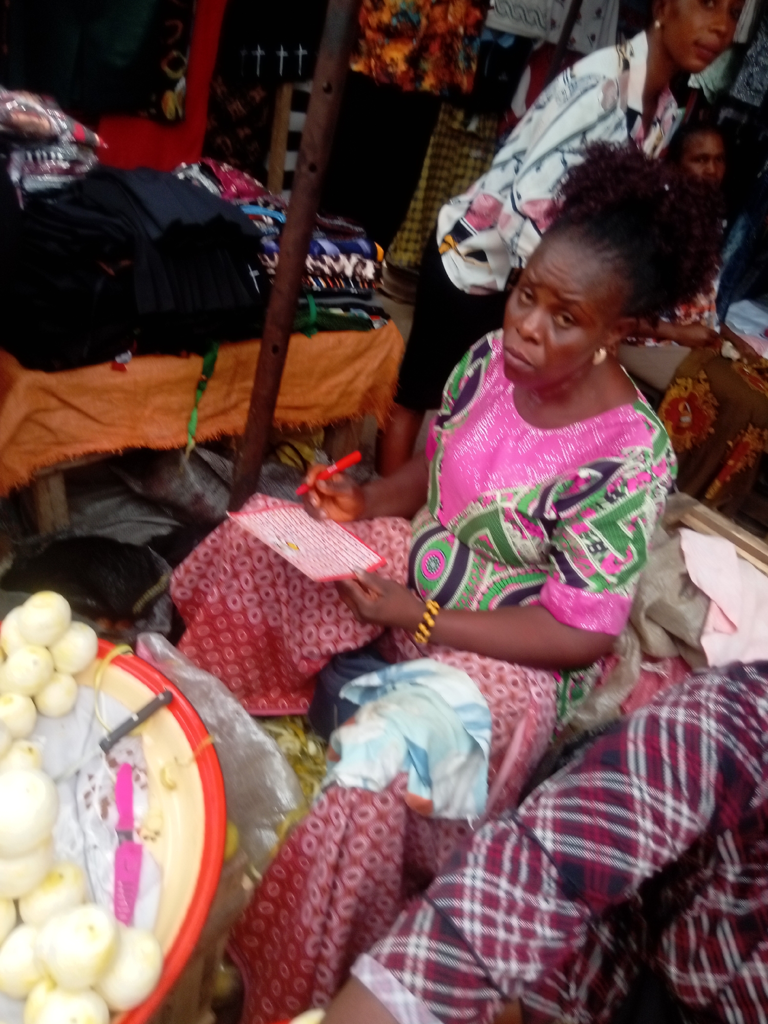In the city of Fegge, Nigeria, an eye-level shot captures a woman sitting amid market activity. She wears a pink, green, and purple patterned dress. In her hands, she holds a piece of paper and a red pen. Next to her, a red-rimmed tray contains white onions. Other people are present in the background. Clothing items are visible on a table behind the woman. The scene suggests a bustling marketplace.