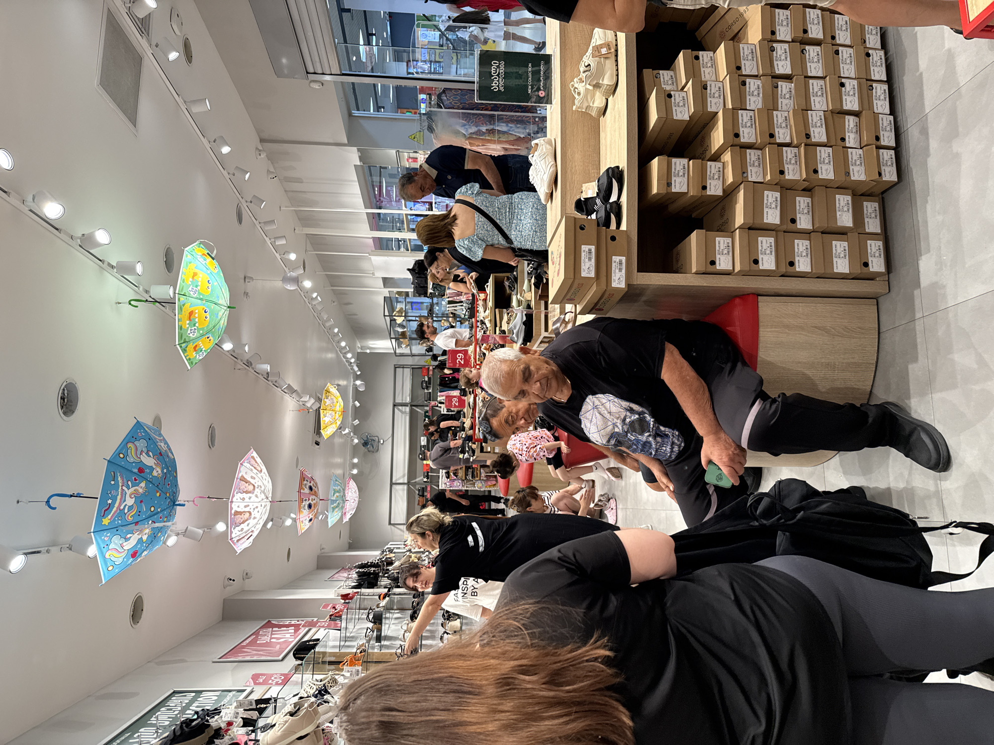 The image shows the interior of a shoe store, likely in a mall, as indicated by the generic store layout and ceiling lighting. Several people are browsing and interacting with the merchandise. In the foreground, a woman with long brown hair is facing away from the camera, wearing a black t-shirt and grey leggings. To her right, another woman in a black t-shirt points towards the ceiling.
Suspended from the ceiling are several brightly colored, patterned umbrellas, acting as decorative elements. Various shoe displays are visible throughout the store, with shelves stocked with boxes and individual shoes. People of different ages are present, some looking at shoes, others sitting on display benches, and some standing and talking. A sign with Georgian text is visible on a display. In the background, a rack of clothing is also present, suggesting the store may sell more than just shoes. The overall atmosphere appears to be a busy retail environment.
