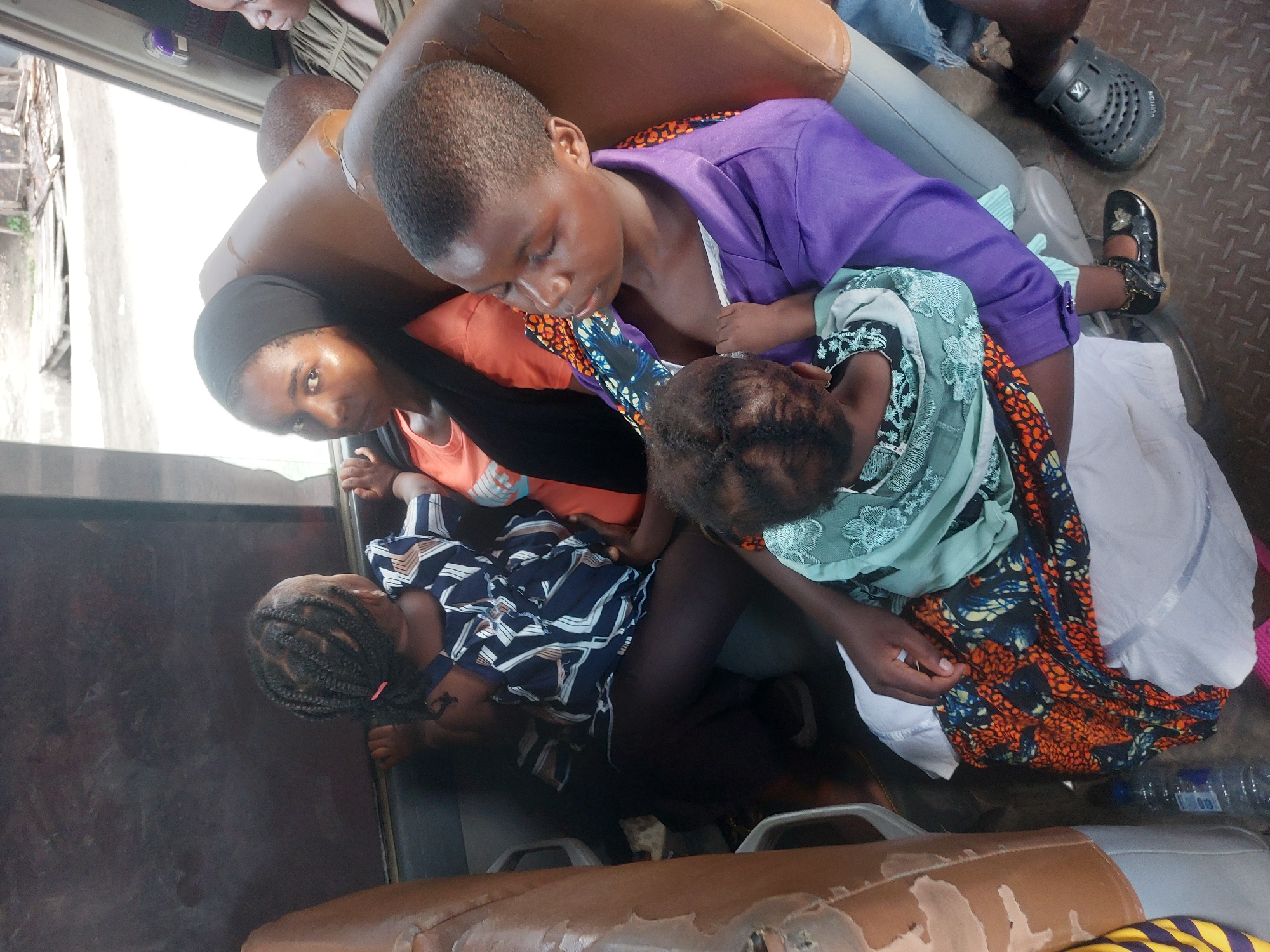 The photograph shows a crowded scene inside what appears to be a public transport vehicle, possibly a daladala, a type of minibus commonly used in Dar es-Salaam, Tanzania. Four individuals are visible, two adults and two children.
An adult woman with a black hijab is looking directly at the camera. She's wearing an orange top. Next to her, a young girl with braided hair is lying on the seat. Another adult, dressed in a purple shirt, holds a child. The child has an orange-patterned cloth. The setting appears to be cramped, typical of public transport in densely populated urban areas. The interior of the vehicle looks worn, with visible signs of use on the seats. A water bottle is also seen in the scene.