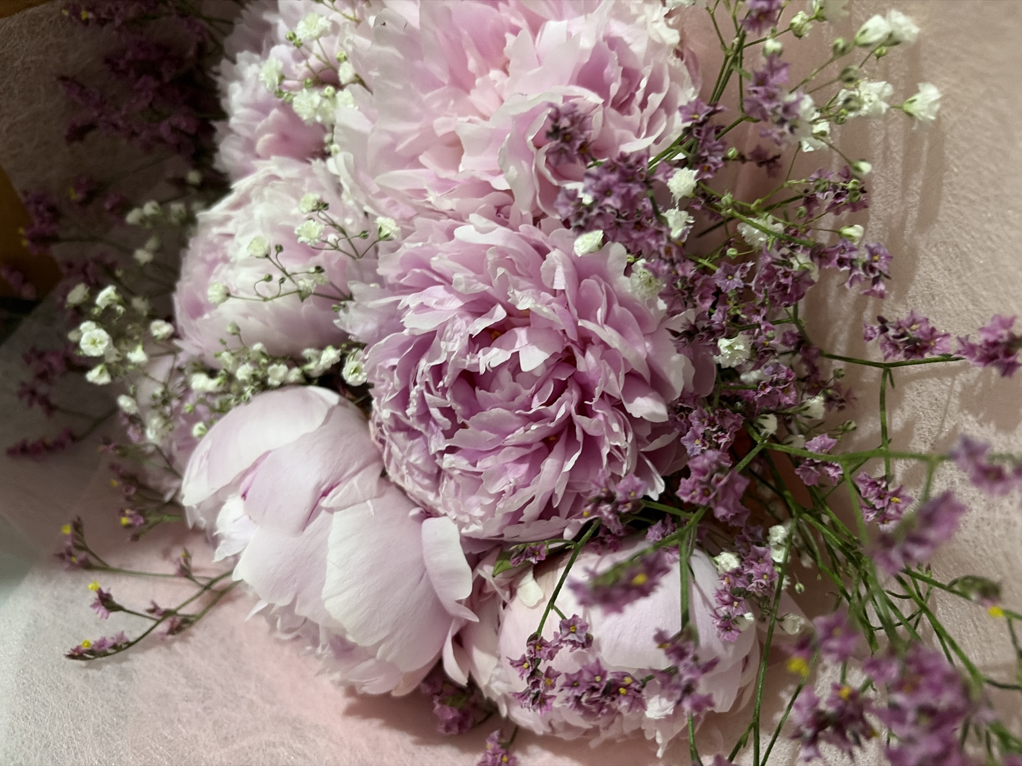 A close-up, overhead shot captures a bouquet of pink peonies and smaller purple and white flowers. The peonies, in various stages of bloom, display layers of delicate petals. Interspersed among the peonies are sprigs of tiny purple flowers with yellow centers and small clusters of white baby's breath. The bouquet is arranged on a soft, textured pink background, possibly fabric. The lighting is soft and diffused, highlighting the textures and colors of the flowers. The scene appears to be indoors.