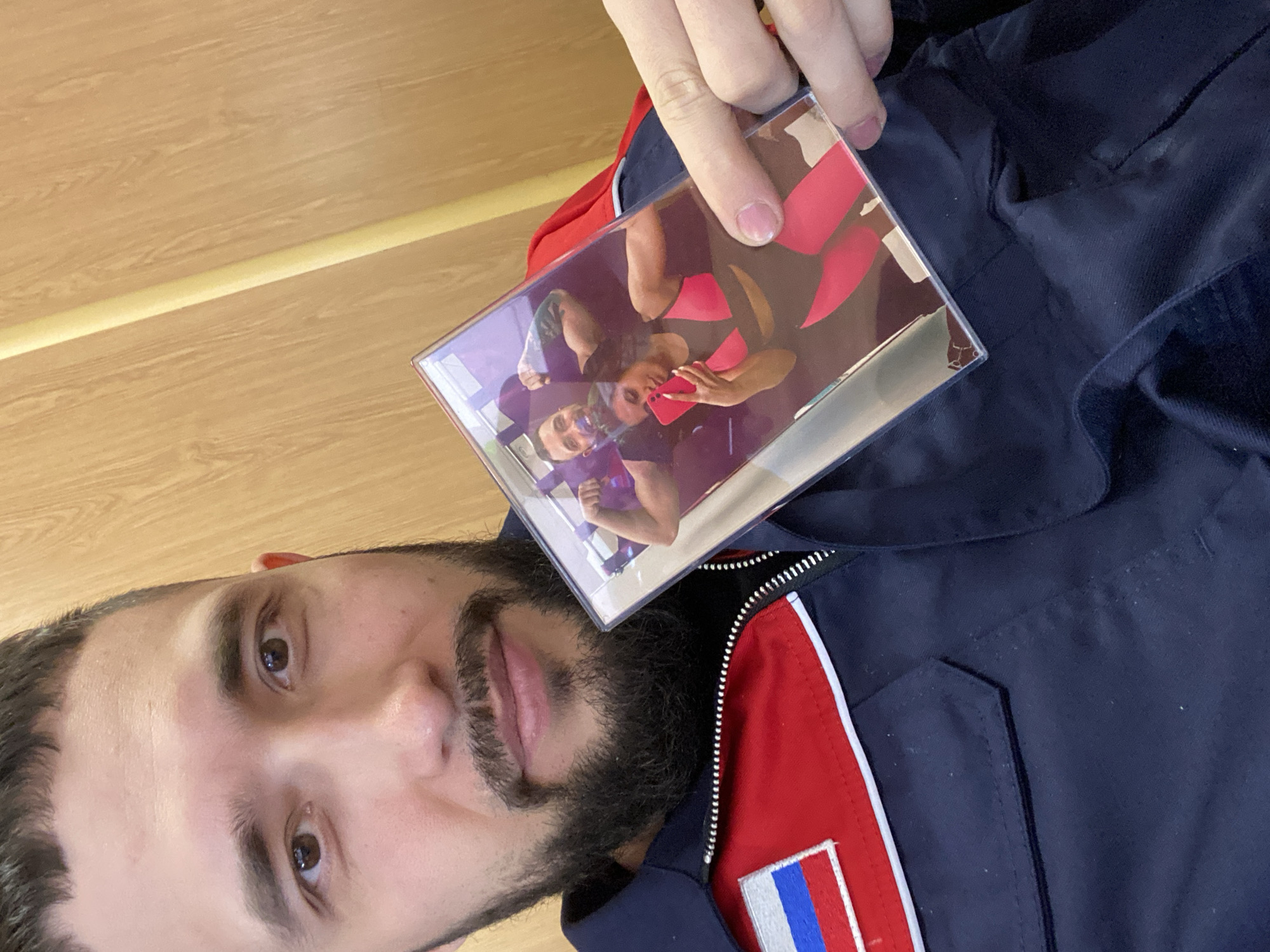 A bearded man is holding a clear, rectangular object containing a photograph or image. He appears to be wearing a dark blue jacket with red and white stripes and a Russian flag patch on the left side. The background shows a light-colored wooden wall. The location is not discernible from the image.
