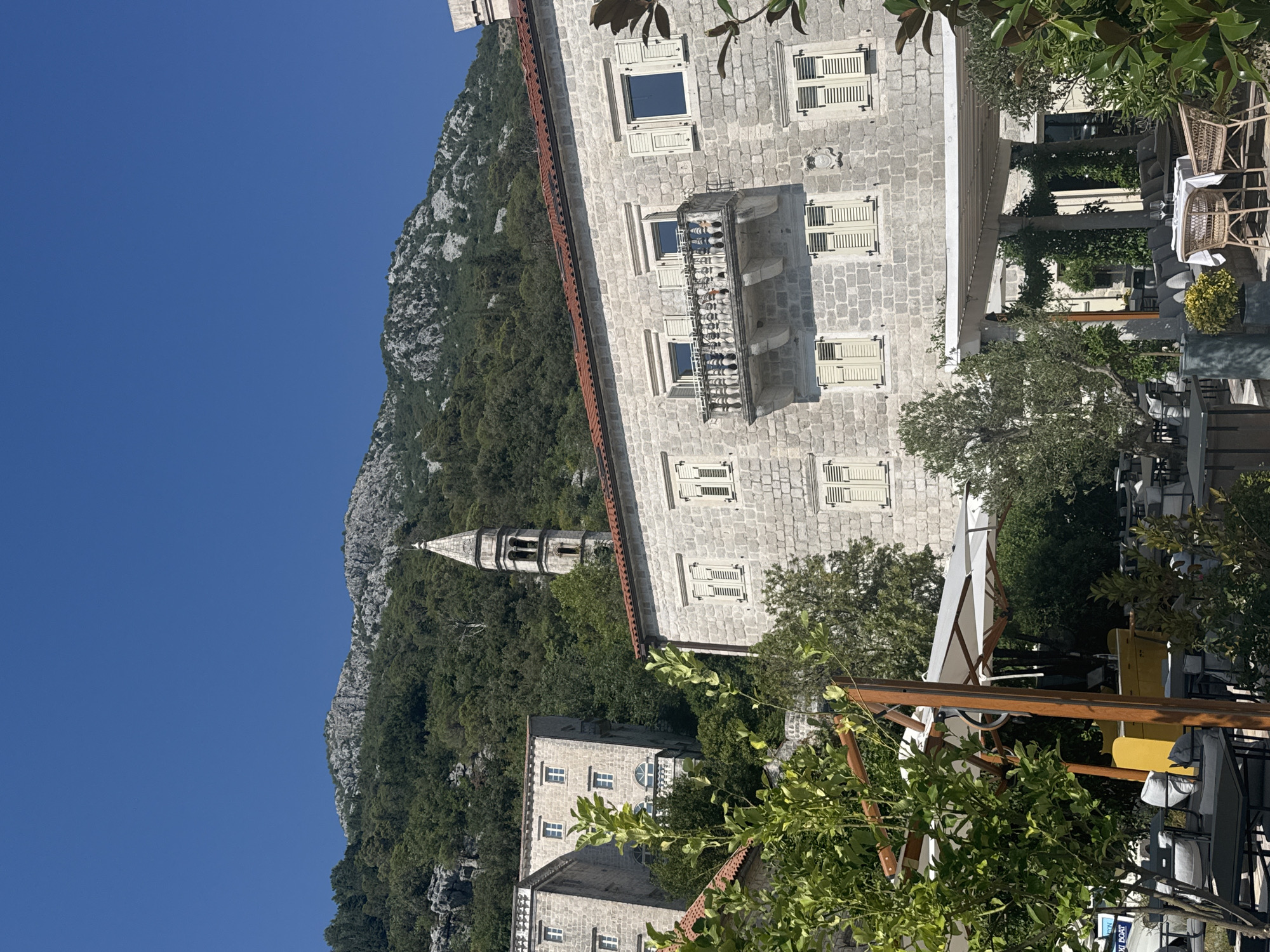 A low-angle shot captures the architecture of Perast, Montenegro, under a clear blue sky. In the foreground, a lush green tree with bright leaves partially obscures a white patio umbrella and outdoor seating area. The umbrella is open, casting a shadow over several tables and chairs. Behind the umbrella, a yellow structure is visible, and more outdoor seating can be seen.
The middle ground features a pale stone building with a red-tiled roof. It has a prominent balcony with ornate white railings. Several windows with light-colored shutters are visible on the building's facade. To the left of this building, another pale stone structure rises higher, also featuring multiple windows with shutters.
In the background, a tall, slender bell tower with arched openings stands amidst dense green foliage. Behind the tower and buildings, a rugged mountain slope covered in trees and rock formations dominates the upper portion of the image. The mountain has a distinct rocky texture and appears arid in some areas. The overall scene suggests a sunny day in a historic European town.