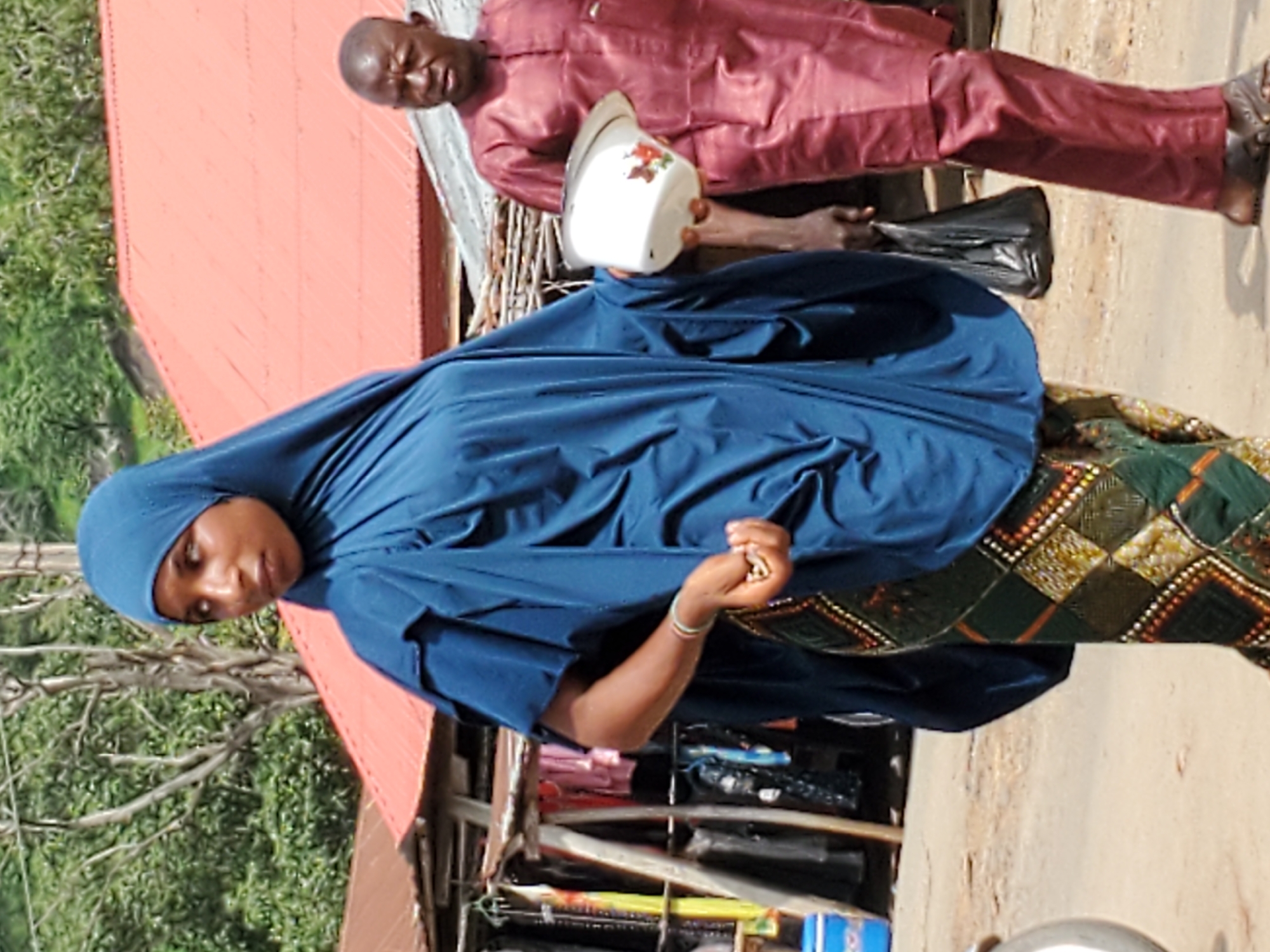 In Mararaba, Nigeria, a woman wearing a dark blue hijab and patterned dress stands on a dirt road. She holds small objects in her right hand. Behind her is a makeshift stall with various merchandise. A man in a maroon outfit stands behind her, holding a white container with floral decorations and a black plastic bag. The background shows a red roofed building and green trees.