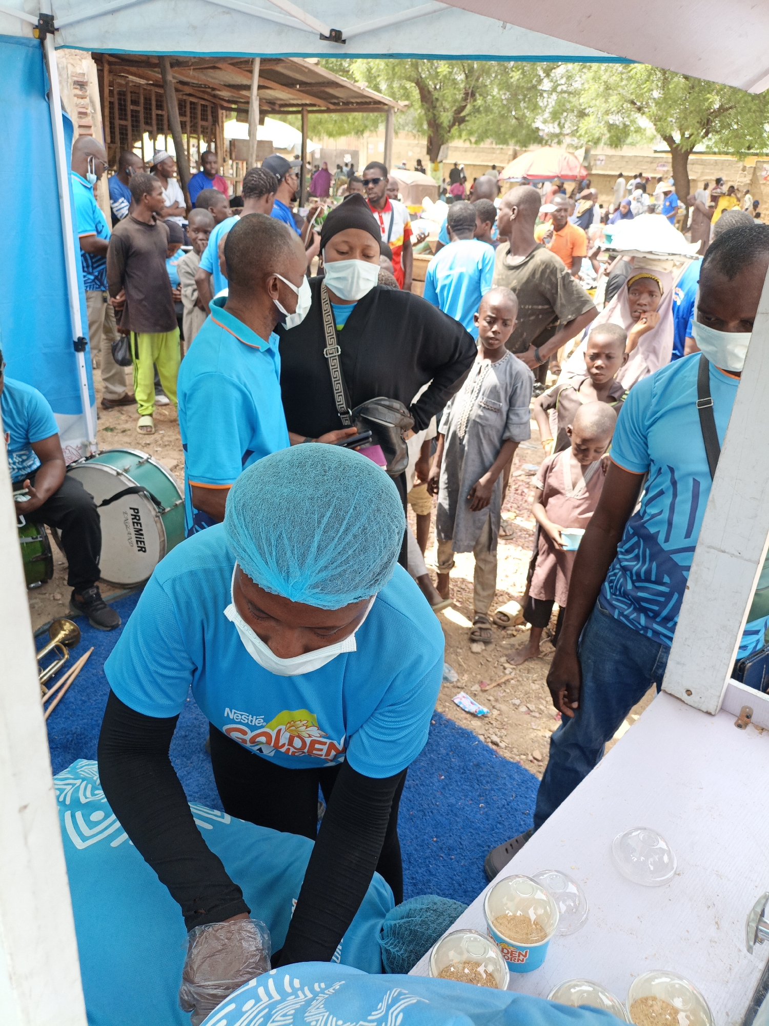 A medium, eye-level outdoor shot captures a scene in Kawaji, Nigeria. A person wearing a light blue shirt, hairnet and gloves, leaning over a white table and preparing what appears to be food samples. The shirt shows the Nestle Golden Morn brand. On the table are small cups filled with a granular substance.
Several individuals stand nearby, observing the scene. Many are wearing face masks. The environment appears to be a busy outdoor market, with numerous people and structures in the background. There's a tent setup where people appear to be offering free samples. The backdrop is characterized by crowded streets and trees. A musical drum is seen in the lower left of the frame.