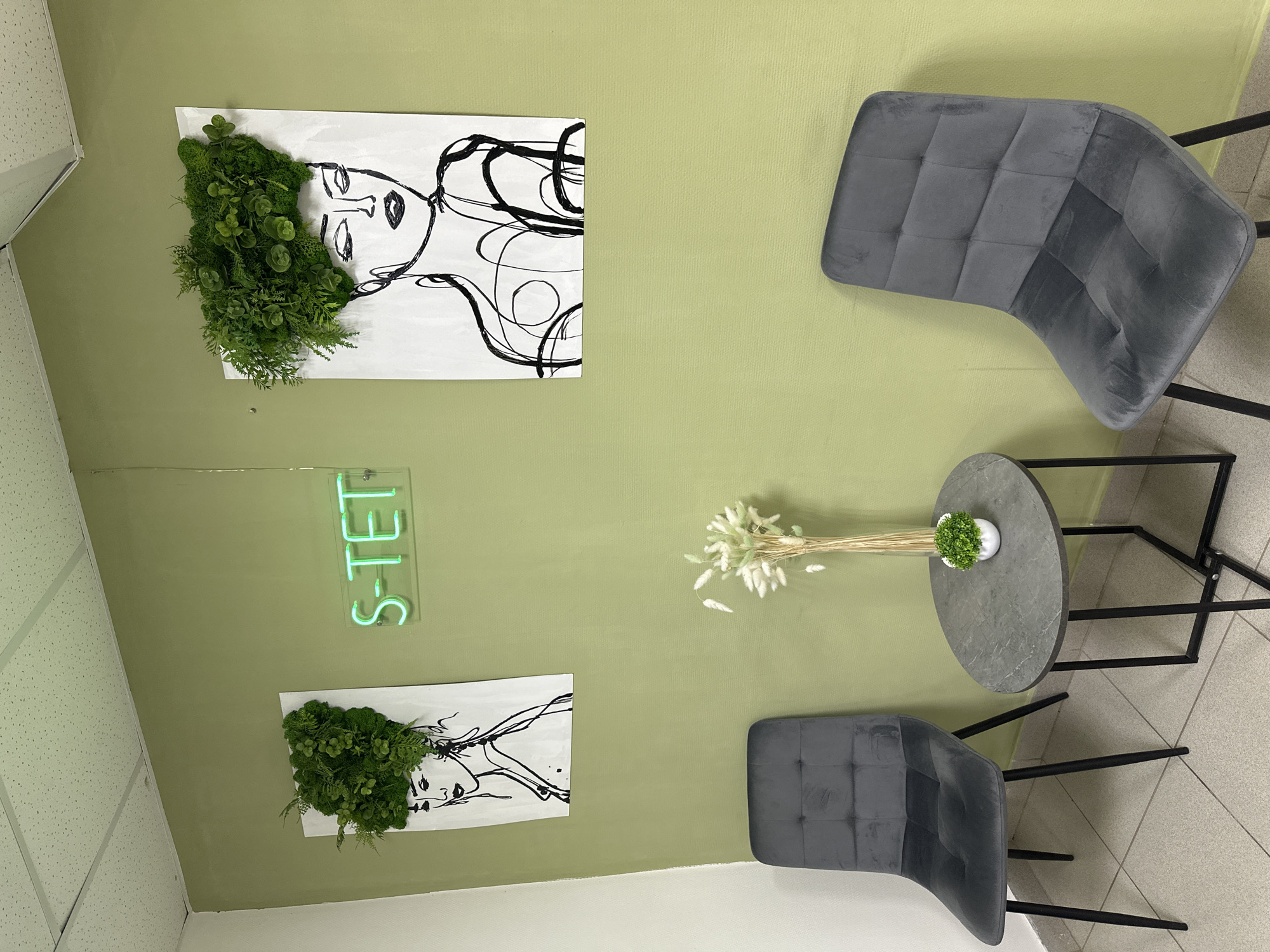 The image displays a clean and modern interior space, likely a waiting area or lounge within a commercial establishment in Chaikovsky, Russia. The dominant feature is a light sage green wall that serves as a backdrop for the room's decor. The flooring consists of light-toned ceramic tiles, and the ceiling features white acoustic panels arranged in a grid pattern.
Two dark grey velvet upholstered chairs with black metal frames are positioned opposite each other, creating a seating arrangement. Between these chairs is a small, round table with a dark grey, possibly marble-effect, top and slender black metal legs. The table is adorned with a small artificial potted plant featuring vibrant green foliage and a bouquet of dried, light-colored fluffy stalks.
The green wall is decorated with two identical rectangular white canvases. Each canvas features a minimalist black line drawing of a human profile, likely a woman, with stylized lines representing hair. Artificial green plants and moss are affixed to the canvases, creating a unique biophilic effect where the greenery forms the "hair" of the drawn figures. Centered between these two art pieces, a bright neon green sign spells out "S-TET" in capitalized sans-serif letters.
No people are visible in the scene, which is static and appears to be illuminated by indoor lighting. The overall aesthetic is contemporary and inviting, suggesting a calm and well-designed environment.