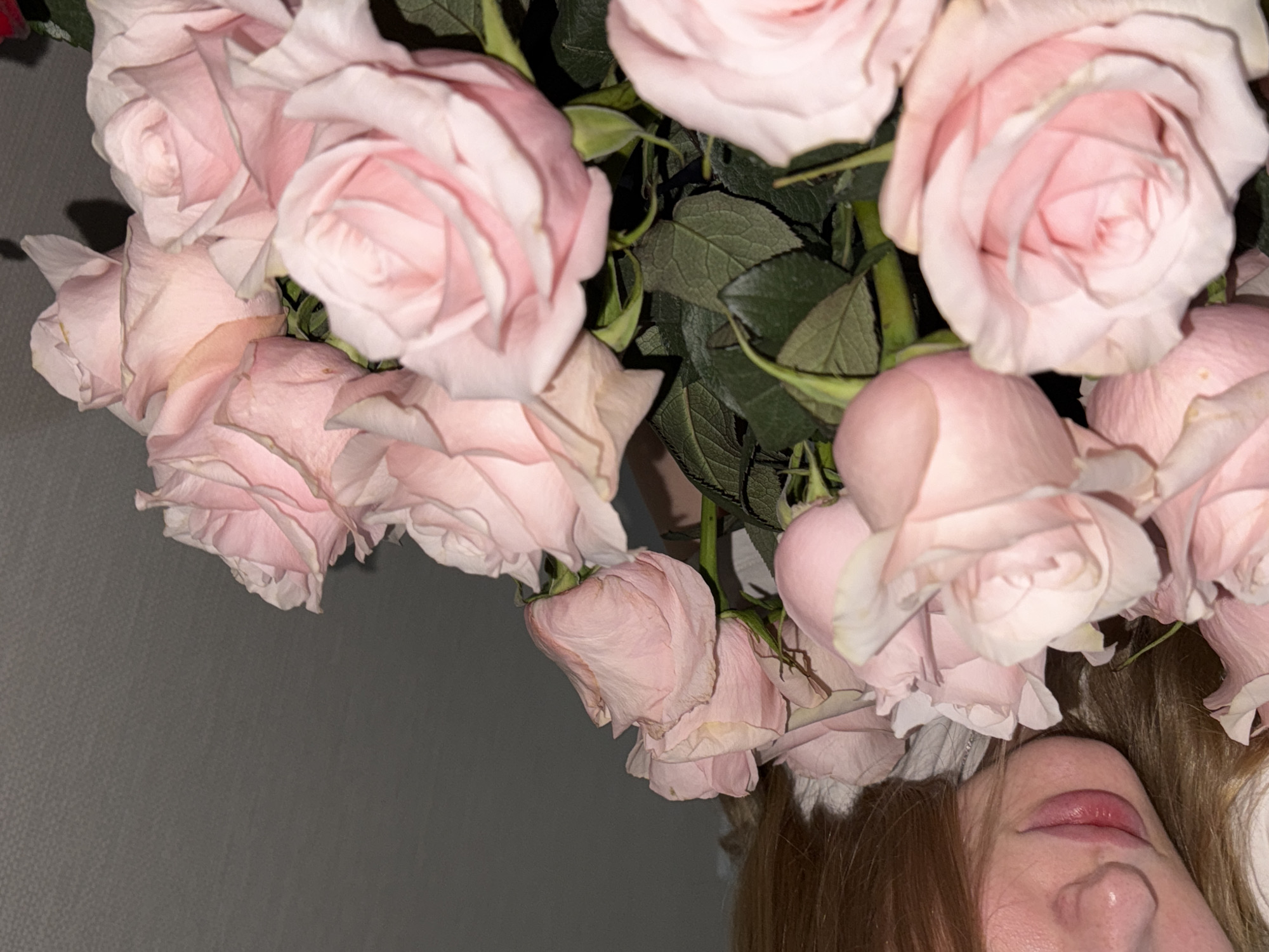 The image shows a close-up of a bouquet of light pink roses. A person's face is partially visible in the upper left corner, with their lips and chin in focus, and their blond hair falling down. The background is a plain grey wall. There are no visible landmarks or contextual clues to suggest a specific location within the city of Perm, Russia. The scene is static and focuses on the flowers and the person's face.