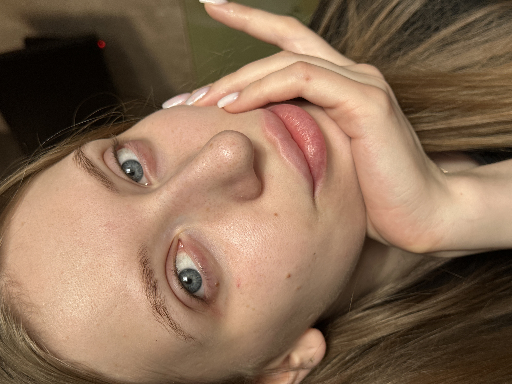 A close-up, tilted portrait of a young woman with light skin, blue eyes, and light brown hair. She is reclining, with her hand near her face. Her nails are manicured with a light color. The background is blurred and consists of dark and light shades, with an indistinguishable object in the upper left corner. There is no discernible context related to the city of Chaikovsky, Russia.