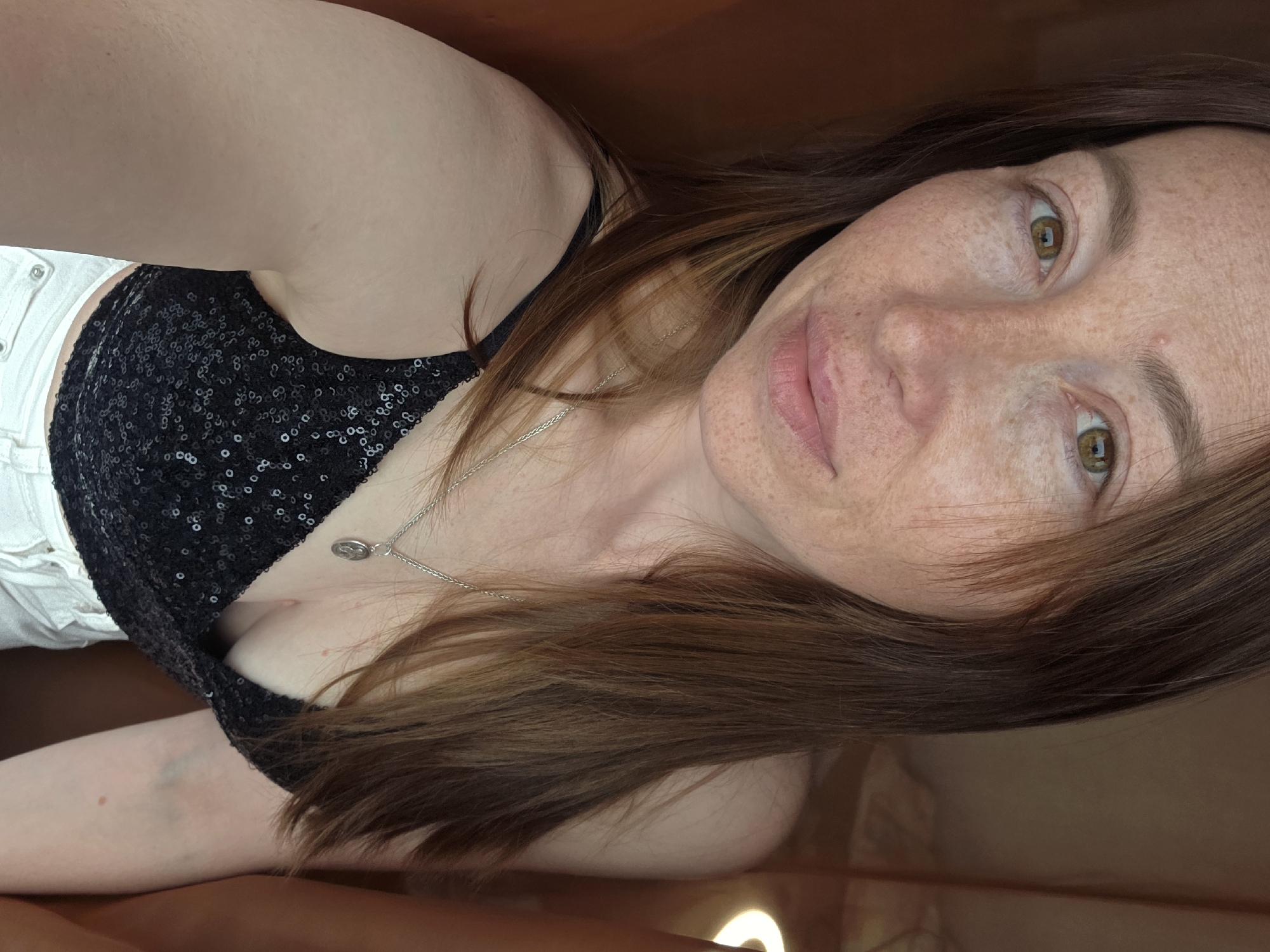A woman with fair skin and freckles poses in an indoor setting. She has hazel eyes and light brown hair that falls across her face and chest. She is wearing a black, sequined top and white shorts. A silver necklace with a circular pendant is visible around her neck. The background appears to be a brown, reflective surface. The photo seems to be taken at an angle, with the woman's head tilted. There is no contextual information within the image to determine a location in the city of Chaikovsky, Russia.