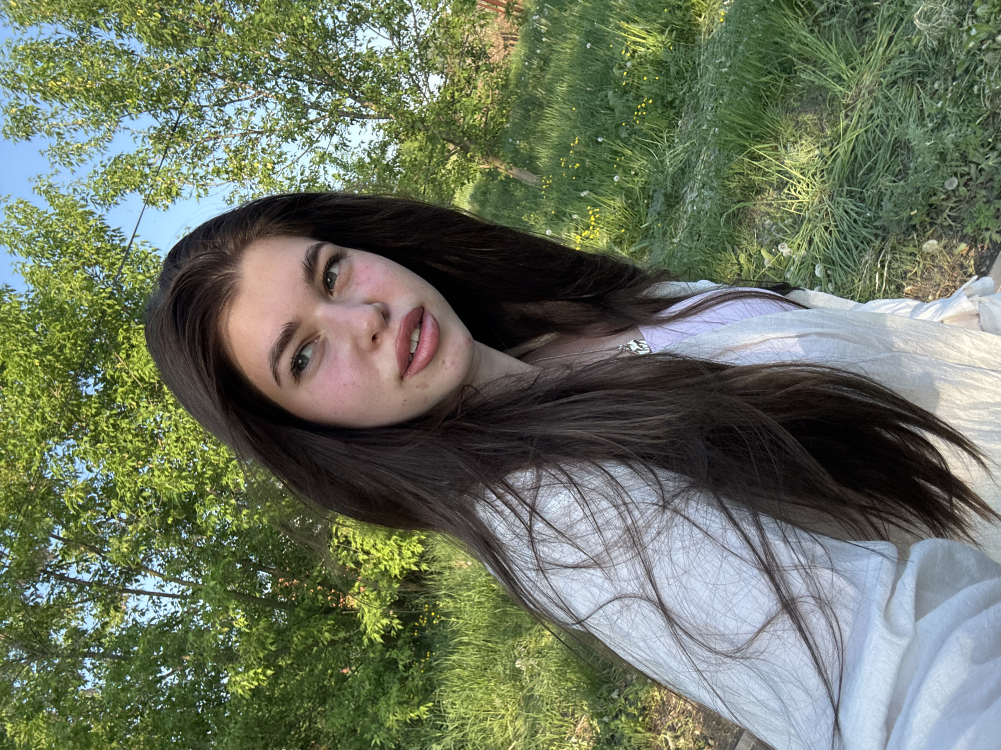 A young woman with long, dark brown hair and fair skin is the subject of this outdoor selfie. She is wearing a light-colored, loose-fitting garment over a lilac top. Her green eyes are visible, and she has a smattering of freckles. The background features lush green grass and a tree with bright green foliage, indicating a natural environment. The presence of dandelions suggests the photo was taken during the warmer months. The angle and composition suggest she is lying or reclining on the ground. The location is Troshkovo, Russia.