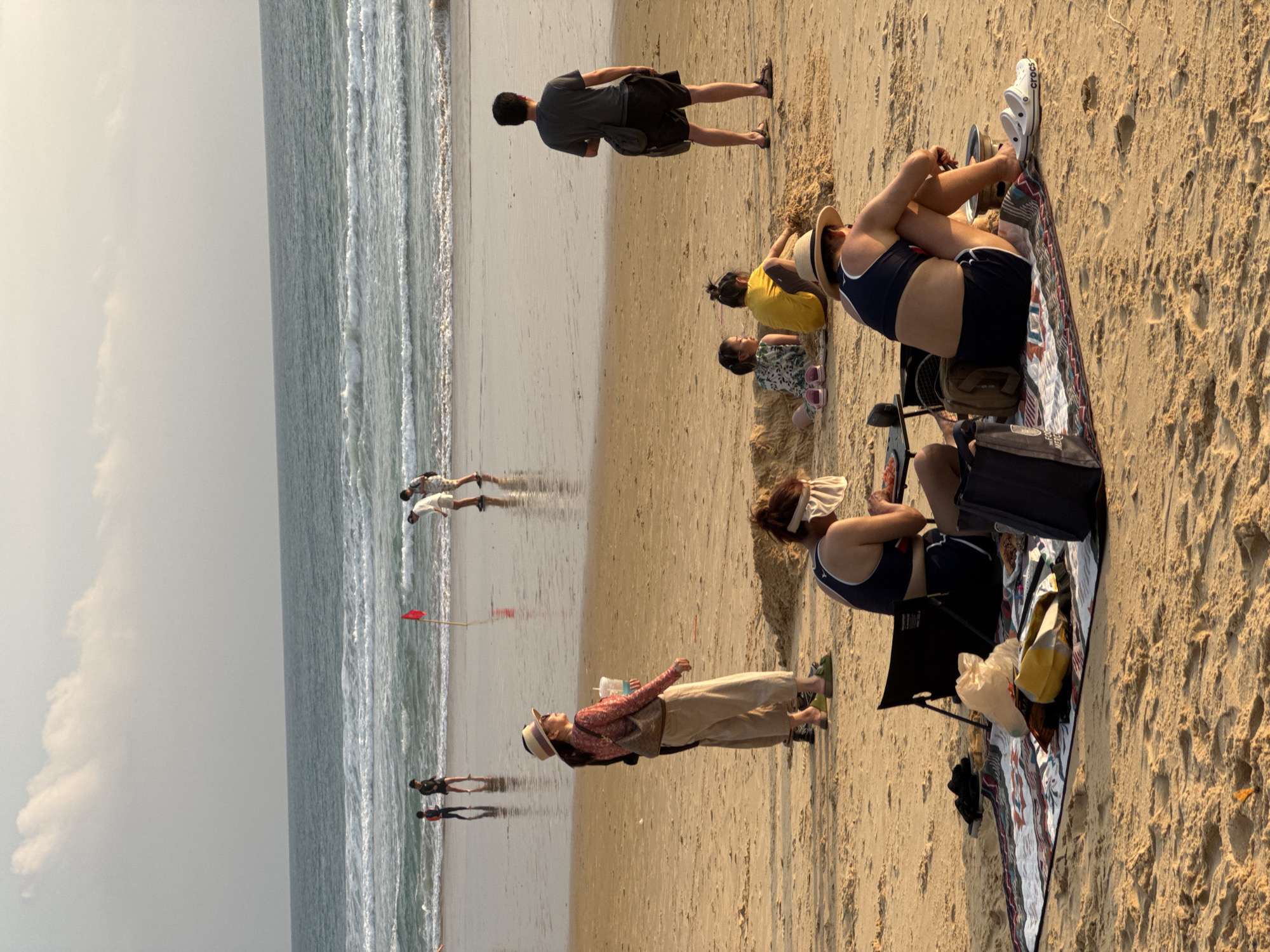 The image depicts Karon Beach in Karon, Thailand, with a clear sky and calm ocean waves. In the foreground, a group of people are enjoying a picnic on a blanket spread on the sand. A woman in a straw hat and blue bikini top is seated, facing away from the camera, with a portable grill and food items nearby. Another person is partially visible next to her. In the mid-ground, a woman in a straw hat and patterned top stands with a drink in hand, looking towards the ocean. Behind her, two children are playing in the sand. Further in the background, several individuals are walking along the shoreline, and a red flag stands on the wet sand. The overall scene conveys a relaxed beach atmosphere.