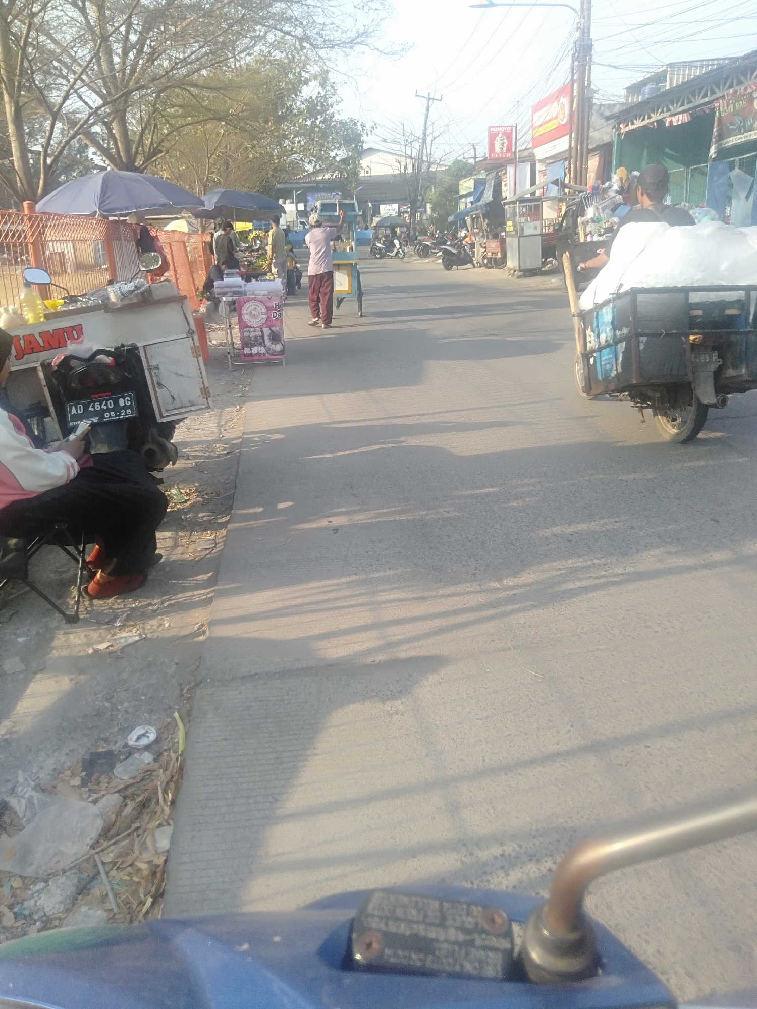 The image captures a street scene in Sukadami, Indonesia, during what appears to be the late afternoon, given the long shadows cast by the sun. The foreground is dominated by the handlebars and front of a blue motorcycle, suggesting the photo was taken from a rider's perspective.
To the left of the motorcycle, a stall is set up with a sign reading "JAMU." A person is seated on a small chair next to this stall, looking at their phone. The stall has a license plate on the back that reads "AD 4640 OG 05-26." A woman with dark hair and light-colored clothing is partially visible behind the stall. Next to the "JAMU" stall is another smaller stall, displaying various items, possibly food or drinks.
Further down the street, a man wearing a light brown long-sleeved shirt and dark pants is pushing a cart filled with white sacks. He is walking on the right side of the road. Behind him, a motorcycle with a trailer is moving away from the camera, also carrying a large white load. Several other motorcycles and a few small carts are visible along the side of the road, indicating a bustling market or commercial area.
The environment is characterized by a paved road with some debris on the side, including plastic bags and fallen leaves. Several trees with bare branches line the street, and power lines are visible overhead. Buildings with awnings and signage are present on the right side of the road, suggesting shops or other businesses. The overall impression is of a typical street in a populated area of Indonesia, with vendors and daily commerce taking place.