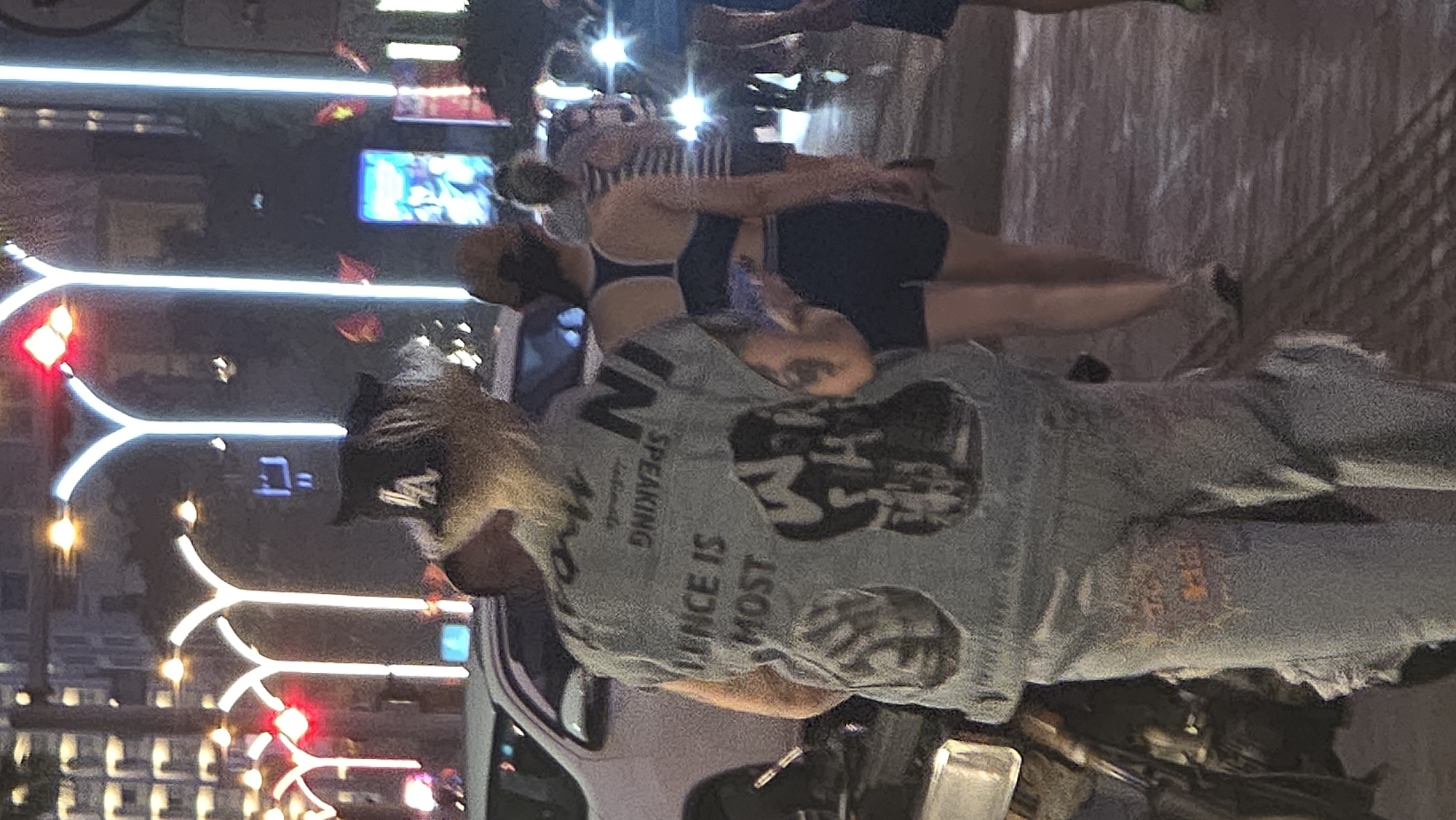 The image shows a street scene at night, likely in Nha Trang, Vietnam, judging by the Vietnamese flags, style of street lights and architecture. A woman wearing a baseball cap and a denim jacket with a graphic on the back stands on the sidewalk. Two women are wearing athletic clothing. The backdrop features illuminated streetlights and buildings. A vehicle is parked nearby.