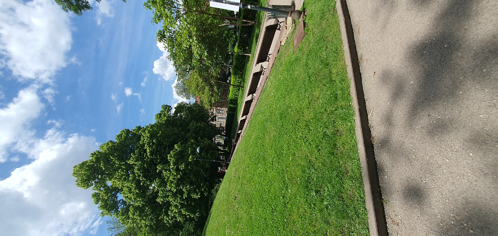 The image depicts a park scene in an urban location. A paved walkway runs along the right edge of the frame. Adjacent to the walkway is a strip of green lawn. Parallel to the lawn is a series of benches facing the opposite direction. In the background, a building is visible through the trees. The sky is blue with scattered clouds. The photograph seems to be taken during the daytime. The location is in the city of Unknown, Unknown.