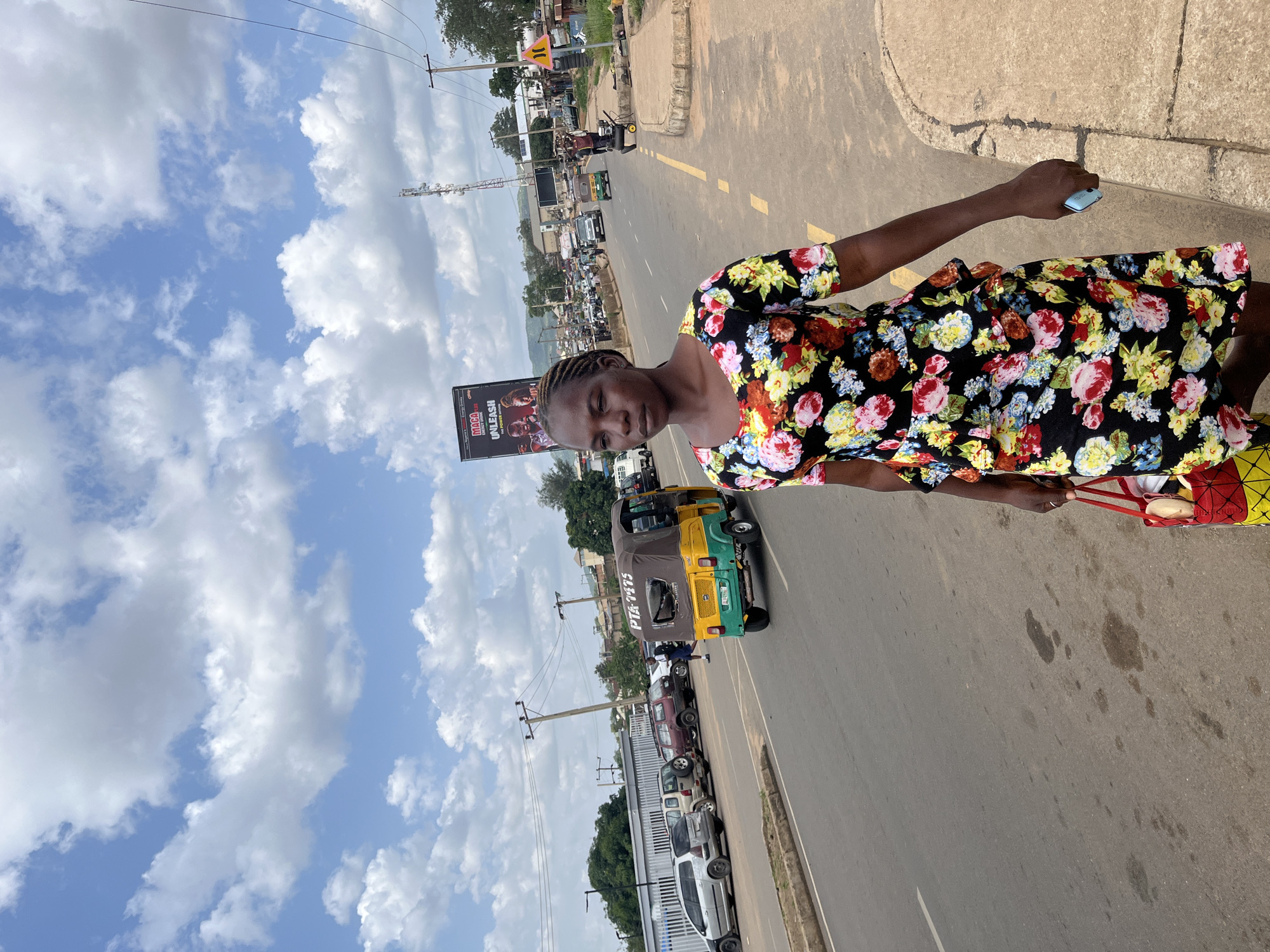 In the city of Rafin Par, Nigeria, a Black woman with braided hair is walking on a paved street with a sidewalk on the right. She is wearing a black dress with a vibrant floral pattern, and carrying a red and yellow patterned bag in her left hand. She holds a light blue smartphone in her right hand. In the background, there is traffic, including a yellow and green auto-rickshaw with the license plate "PTA 7475" and several cars parked on the left side of the road. A large billboard with an advertisement for "MACA UNLEASH" is visible further down the street, along with cell towers and utility poles. The sky is partly cloudy with white cumulus clouds against a blue sky.